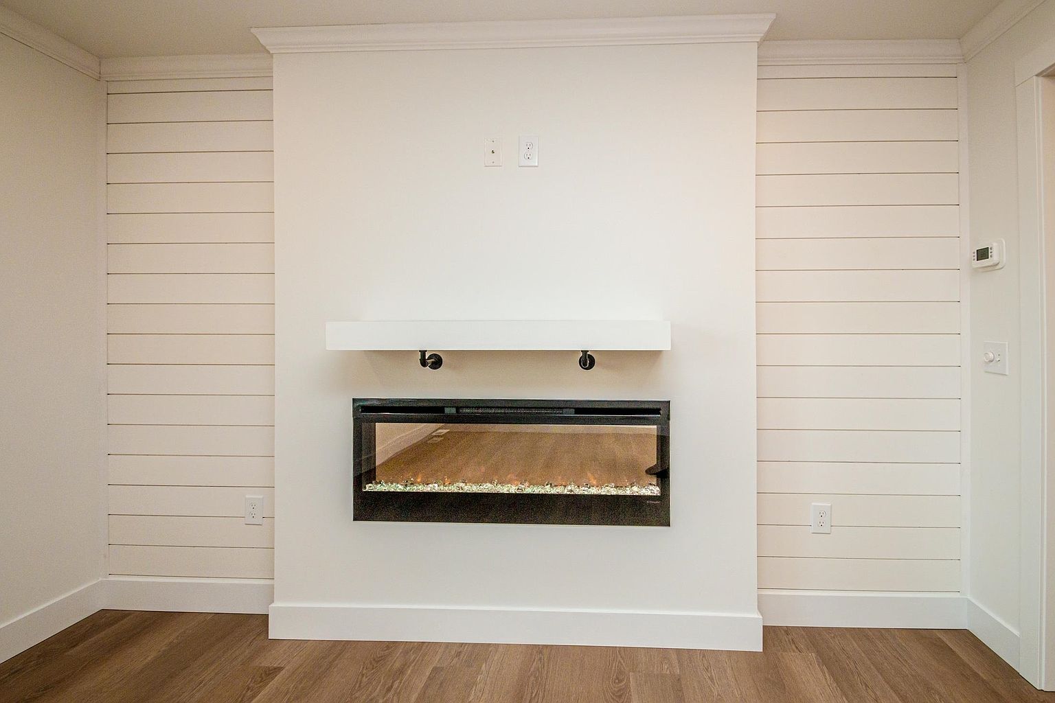 This interior shot features a clean, modern living room focal point centered around a sleek, wall-mounted electric fireplace with a minimalist white mantel. The fireplace is flanked by white shiplap wall paneling, creating a bright and contemporary aesthetic that contrasts with the warm-toned wood flooring. The composition is symmetrical and balanced, highlighting the architectural detail of the fireplace wall as a primary design feature.