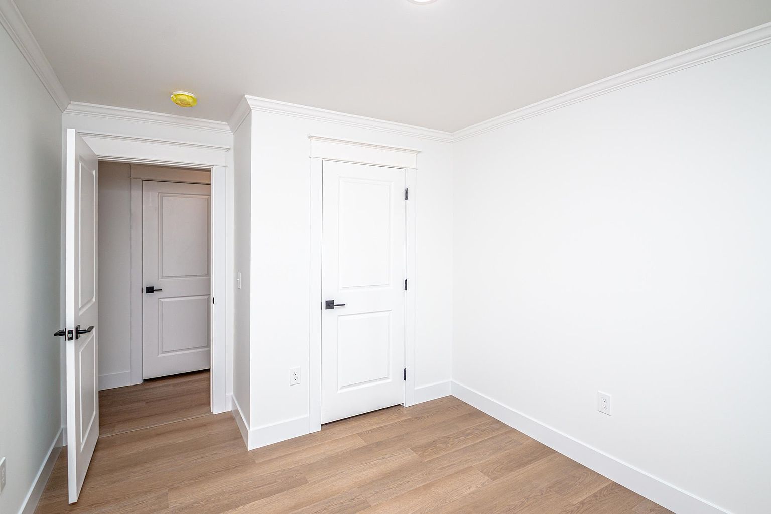 This interior view captures a clean, minimalist hallway featuring light wood-tone flooring and crisp white walls. The space includes a partially open door leading to another room and a closed door, both accented with modern matte black hardware. The perspective is eye-level, emphasizing the bright, neutral aesthetic and the architectural simplicity of the home's transition areas.