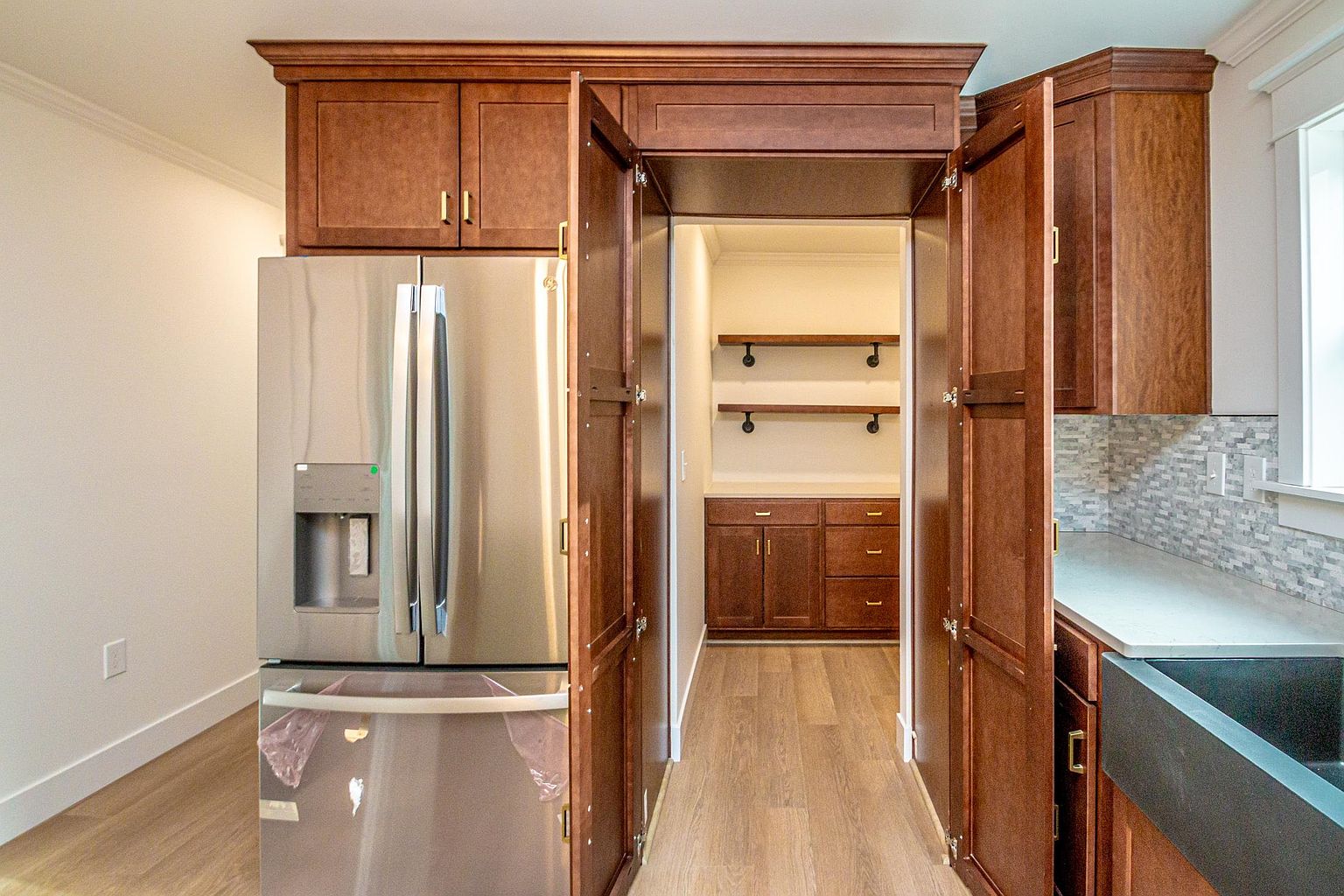 This kitchen features a unique design with a walk-in pantry hidden behind custom cabinetry doors that match the surrounding wood finish. A stainless steel refrigerator is integrated into the cabinetry, while a farmhouse-style sink and light-colored countertops provide a modern contrast to the warm wood tones. The perspective looks directly into the pantry, which is equipped with open shelving and additional lower cabinets, creating a seamless and organized transition between spaces.