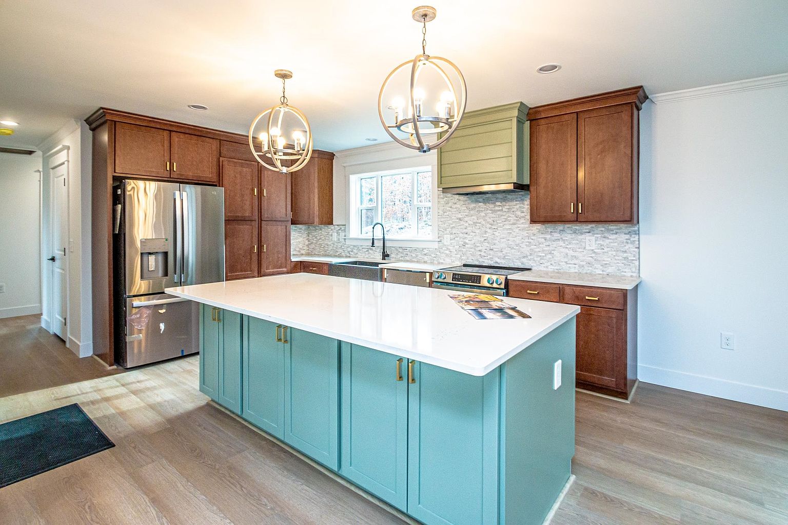 This modern kitchen features a striking contrast between rich, dark wood cabinetry and a vibrant teal-colored center island topped with a pristine white quartz countertop. The space is illuminated by two elegant, spherical pendant lights and includes stainless steel appliances, a tiled backsplash, and a custom-built range hood. The open-concept design and light wood flooring create a bright, inviting atmosphere perfect for both cooking and entertaining.