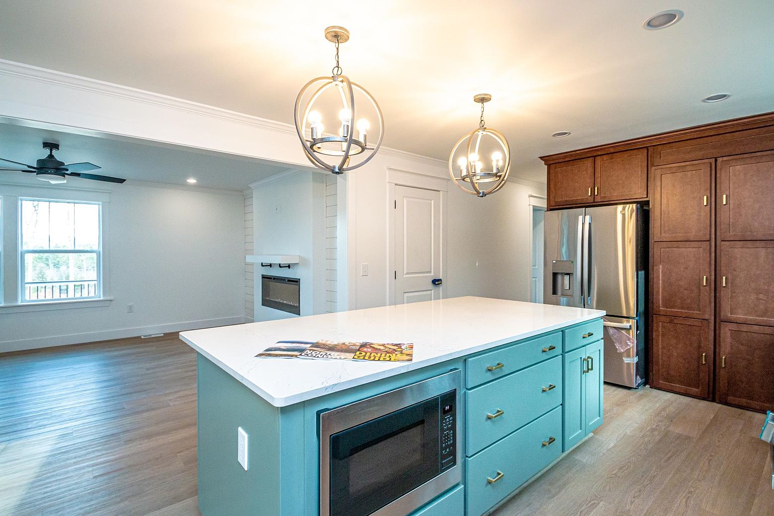 This bright and modern kitchen features a striking teal-colored island with a built-in microwave and white quartz countertops, serving as the focal point of the room. The space is illuminated by two elegant spherical pendant lights and includes warm wood-toned cabinetry paired with stainless steel appliances. The open-concept design flows seamlessly into an adjacent living area, creating an inviting and airy atmosphere perfect for entertaining.