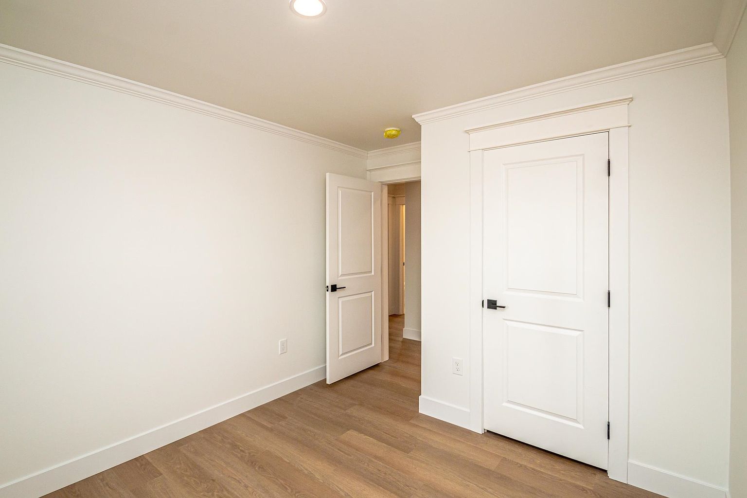 This interior view captures a clean, minimalist hallway featuring light wood-look flooring and crisp white walls. The space includes two white paneled doors, one of which is slightly ajar, leading into another section of the home. The perspective is eye-level and centered, emphasizing the bright, neutral aesthetic and modern architectural finishes.