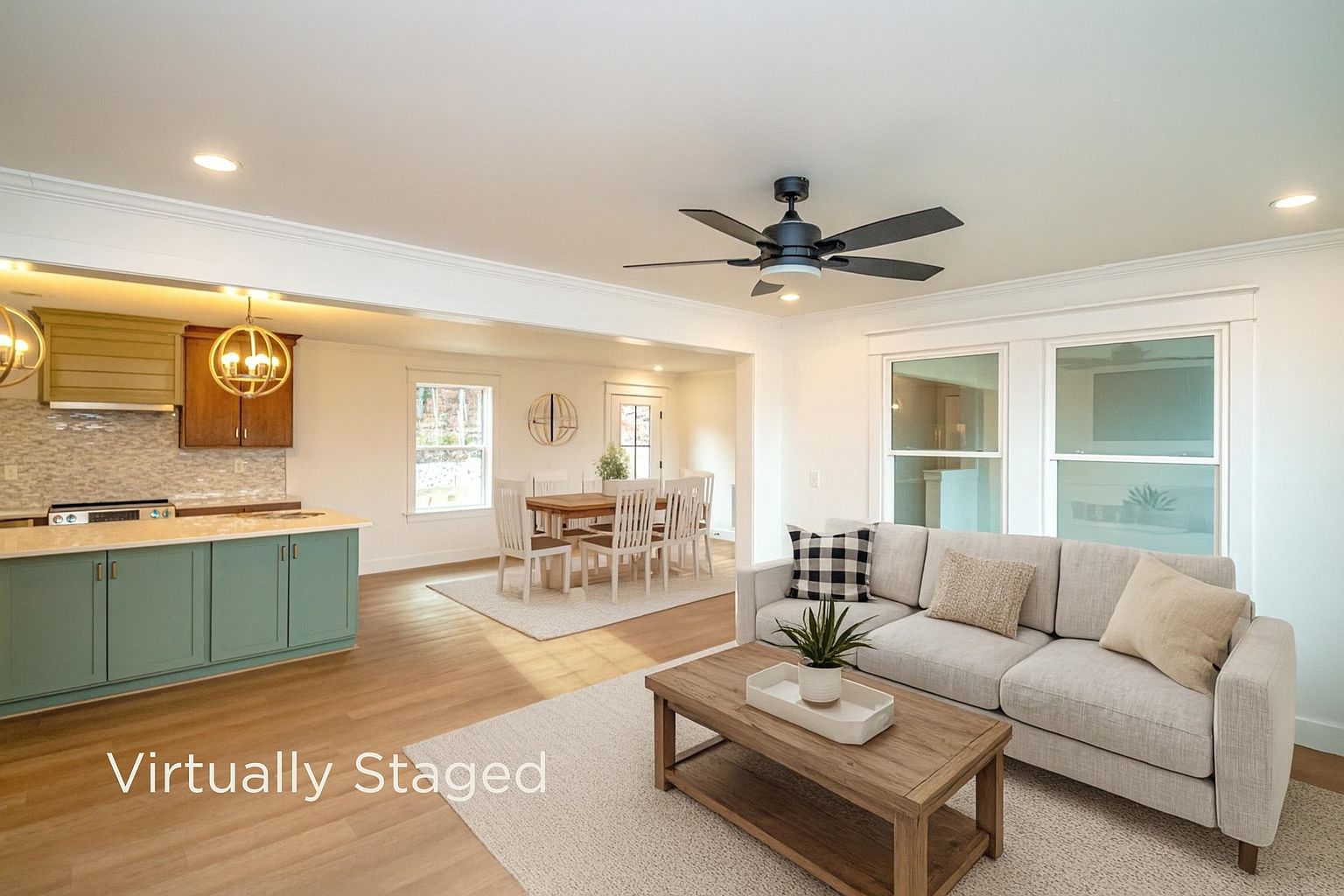 This open-concept living space features a light-filled living room that flows seamlessly into a dining area and kitchen. The room is anchored by a neutral-toned sofa, a rustic wooden coffee table, and a modern black ceiling fan, all set against a backdrop of light hardwood floors and crisp white walls. The virtual staging creates a warm, inviting atmosphere that highlights the home's spacious and airy layout.