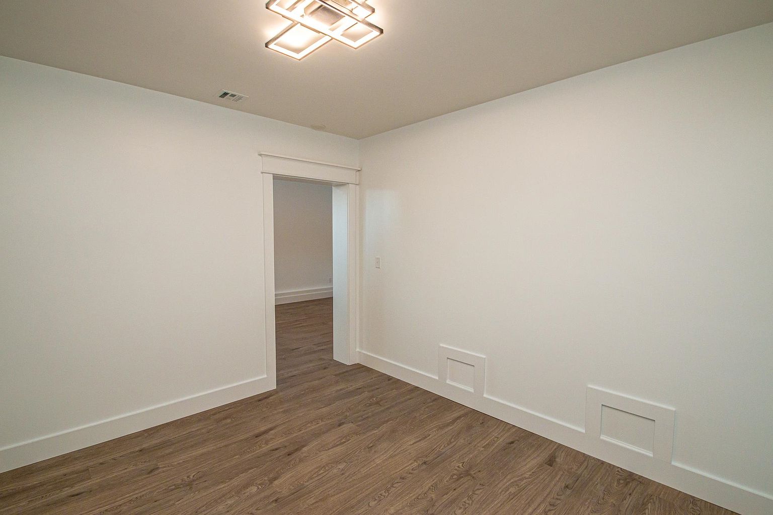 This minimalist bedroom features crisp white walls and warm-toned wood-look flooring, creating a bright and airy atmosphere. A modern, geometric flush-mount light fixture adds a contemporary touch to the ceiling, while decorative wainscoting panels along the lower wall provide subtle architectural interest. The perspective looks directly into the room, highlighting the clean lines and the open doorway leading to an adjacent space.