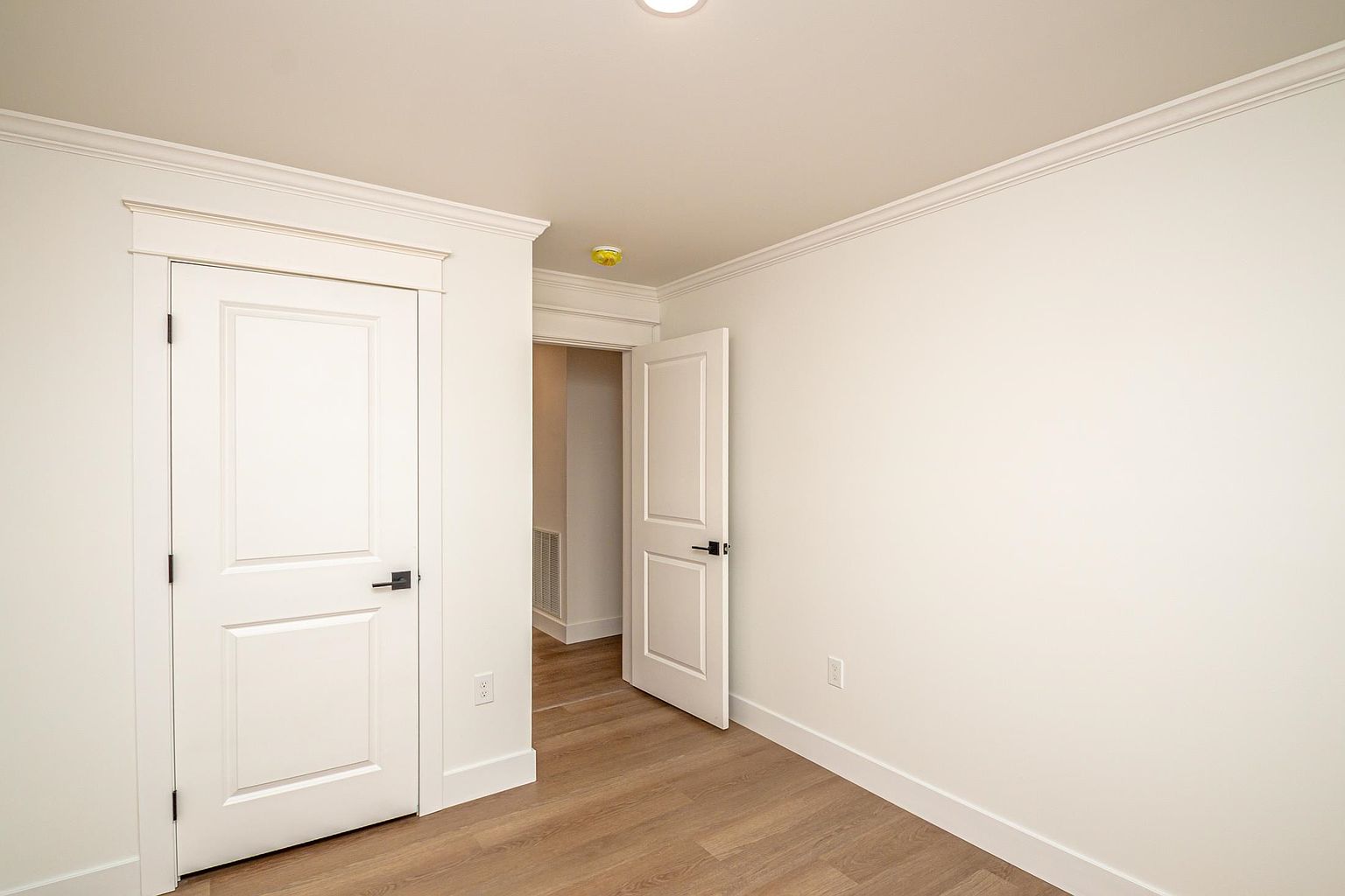 This interior view captures a clean, minimalist hallway featuring light wood-look flooring and crisp white walls. The space includes two white paneled doors with modern matte black hardware, one of which is slightly ajar, leading into an adjacent room. The perspective is eye-level, emphasizing the bright, neutral aesthetic and the architectural detail of the crown molding and door casings.