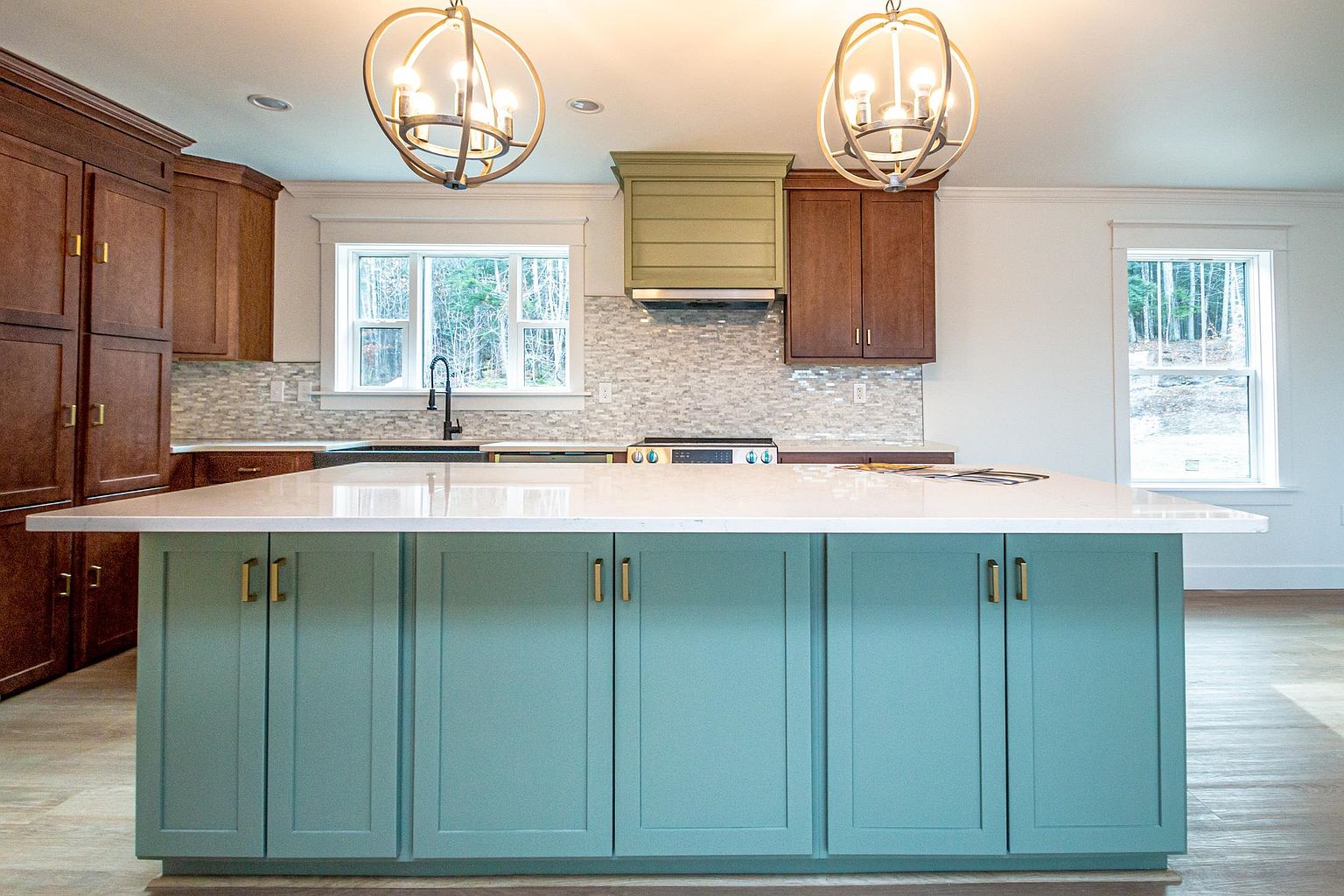 This bright and modern kitchen features a striking teal-colored central island with a white quartz countertop, serving as the focal point of the room. The space is accented by warm wood-toned cabinetry, a unique olive-green range hood, and two large, spherical pendant lights that add a touch of elegance. The perspective is a straight-on, eye-level shot that highlights the clean lines, functional layout, and the contrast between the colorful island and the natural wood elements.