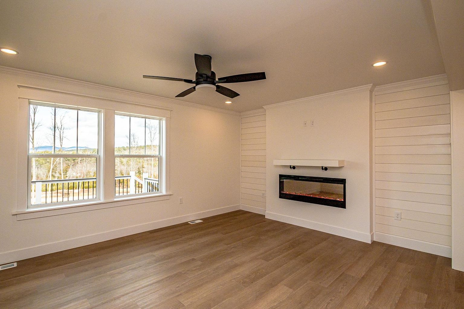This bright and airy living room features modern farmhouse aesthetics, highlighted by white shiplap accent walls and a sleek, wall-mounted electric fireplace. The space is anchored by warm-toned wood-look flooring and a contemporary ceiling fan, while large windows provide ample natural light and a view of the surrounding landscape. The clean, minimalist design creates a welcoming and versatile atmosphere suitable for various decor styles.