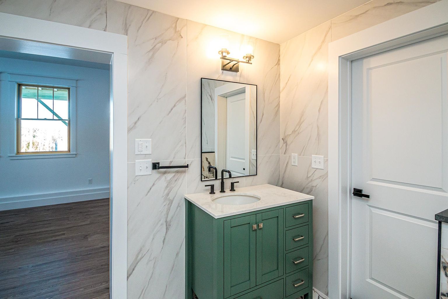 This modern bathroom features a striking green vanity with a white countertop and a sleek, black-framed mirror. The walls are adorned with elegant marble-look tile, creating a sophisticated and clean aesthetic. The perspective captures the vanity as the focal point, with a glimpse into an adjacent room through the doorway.