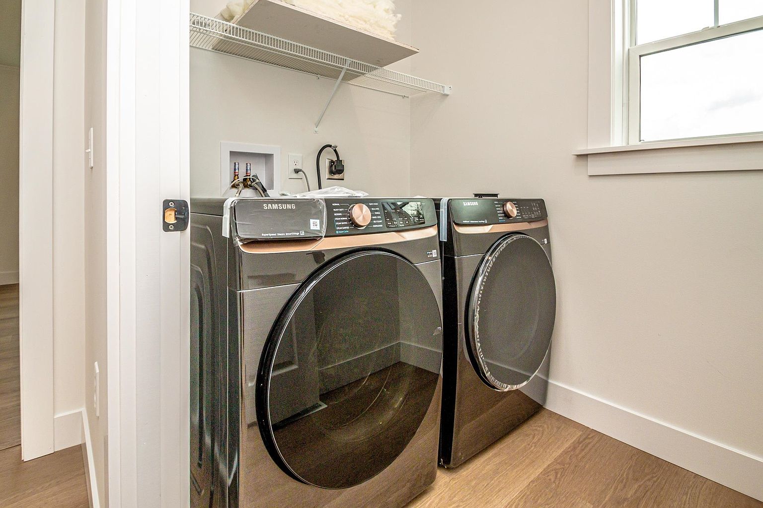 This clean and modern laundry room features a pair of sleek, dark metallic Samsung washer and dryer units positioned side-by-side. Above the appliances, a white wire shelving unit provides convenient storage space, while the room is finished with light-colored walls and wood-look flooring. A window to the right allows natural light to brighten the space, creating a functional and contemporary utility area.