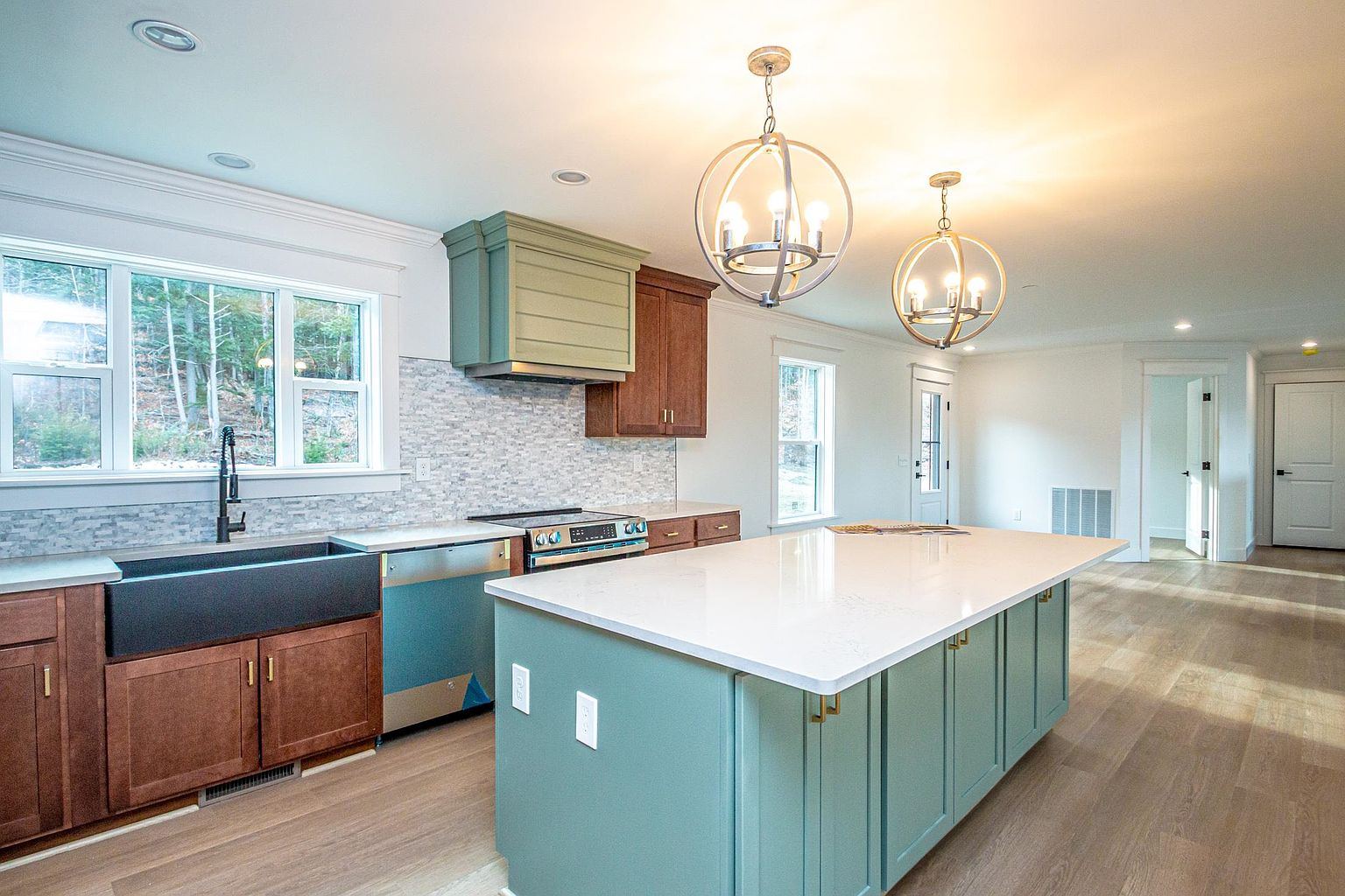 This bright and modern kitchen features a striking contrast between warm wood-toned lower cabinets and a soft sage green island and range hood. A large white quartz-topped island serves as the centerpiece, illuminated by two elegant spherical pendant lights, while a farmhouse sink and subway tile backsplash add a touch of classic charm. The open-concept design flows seamlessly into the adjacent living area, creating an inviting and spacious atmosphere perfect for entertaining.