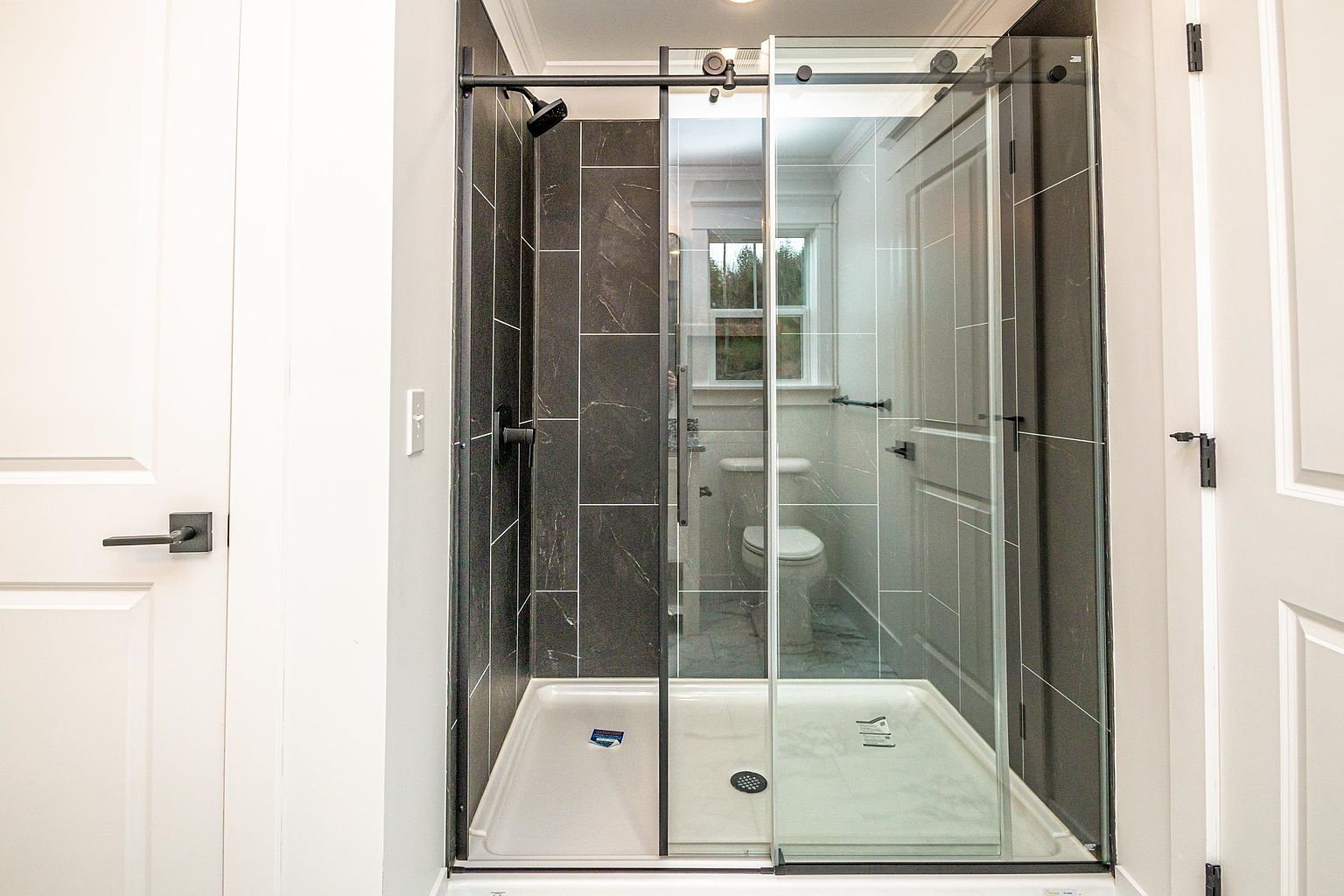 This modern bathroom features a sleek walk-in shower with dark, large-format marble-look tiles and a sliding glass door with matte black hardware. The shower is positioned between two white doors, creating a clean, contemporary aesthetic that maximizes space. The bright, neutral lighting highlights the contrast between the dark shower surround and the surrounding white walls.