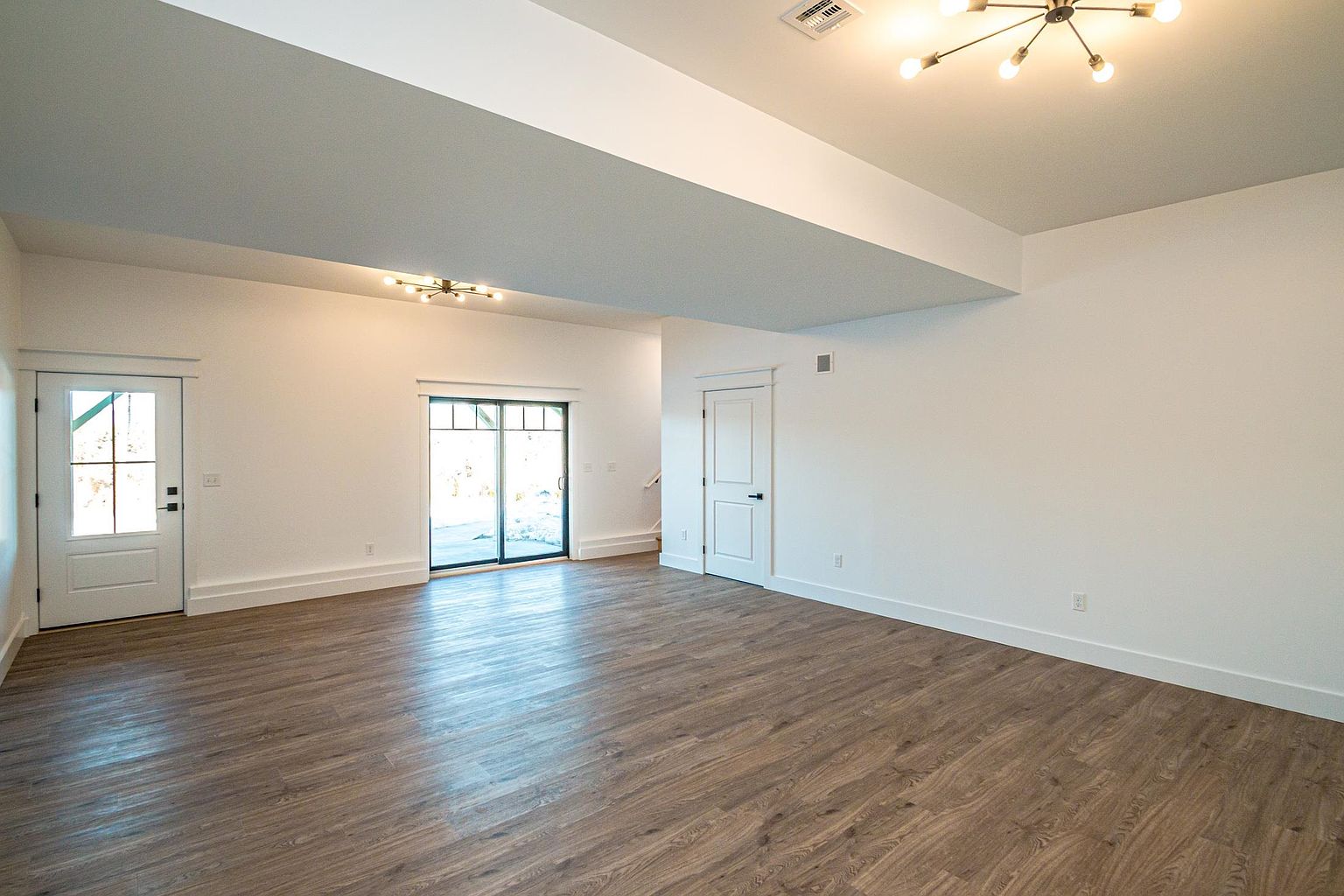 This spacious, open-concept living area features light-toned wood-look flooring and crisp white walls, creating a bright and airy atmosphere. The room is illuminated by two modern, multi-arm sputnik-style chandeliers and includes a glass sliding door that provides natural light and access to the outdoors. The clean lines and neutral palette offer a versatile blank canvas suitable for various interior design styles.