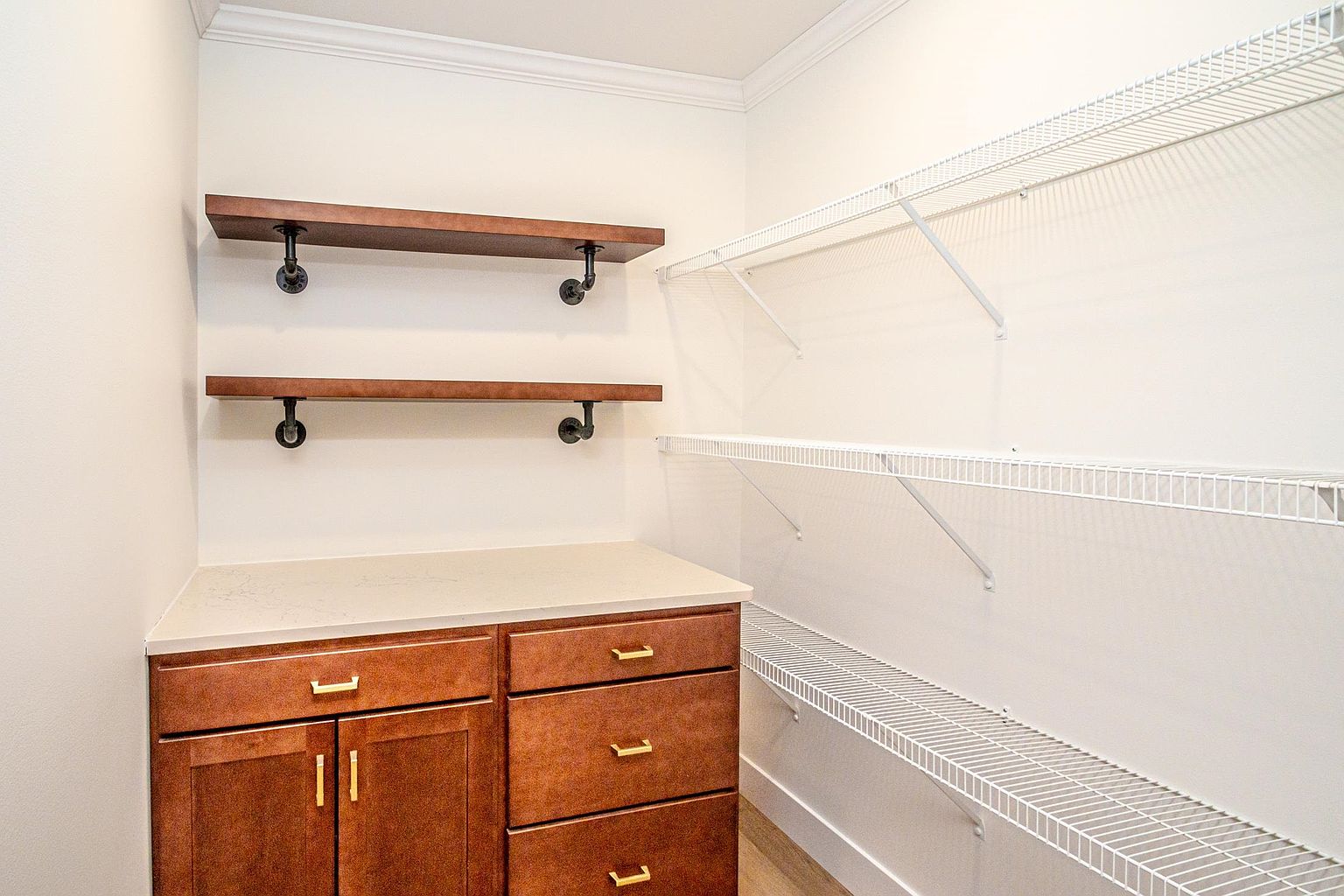 This organized pantry features a combination of custom wooden cabinetry with a countertop and industrial-style floating shelves on the left wall. The right side is equipped with white wire shelving, providing ample storage space for dry goods and kitchen essentials. The clean, neutral aesthetic and functional layout make this an efficient and stylish storage area.