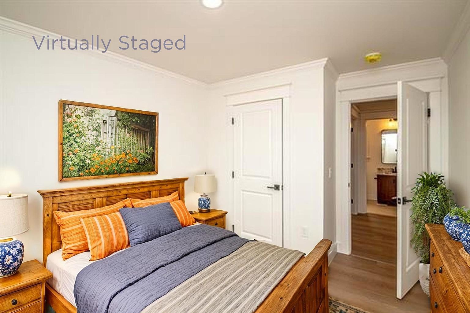 This cozy bedroom features a warm, rustic wooden bed frame dressed in layered orange and blue bedding, complemented by matching wooden nightstands and lamps. A framed landscape painting hangs above the headboard, while an open door reveals a glimpse into an adjacent bathroom with wood-look flooring. The room is bright and inviting, showcasing a clean, traditional aesthetic suitable for a guest space.