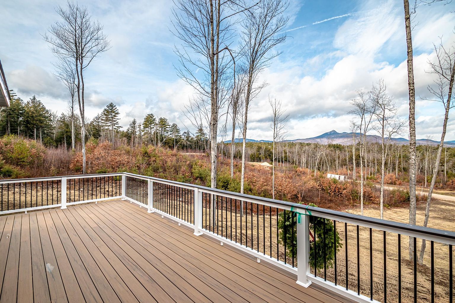 This image showcases a spacious, elevated composite deck featuring clean white railings with black metal balusters, offering a panoramic view of a wooded landscape and distant mountains. The deck is constructed with warm-toned, wood-grain planks and includes a festive holiday wreath attached to the railing, suggesting a cozy, inviting atmosphere. The perspective is from the deck surface, looking out toward the horizon, emphasizing the property's scenic and peaceful outdoor living space.