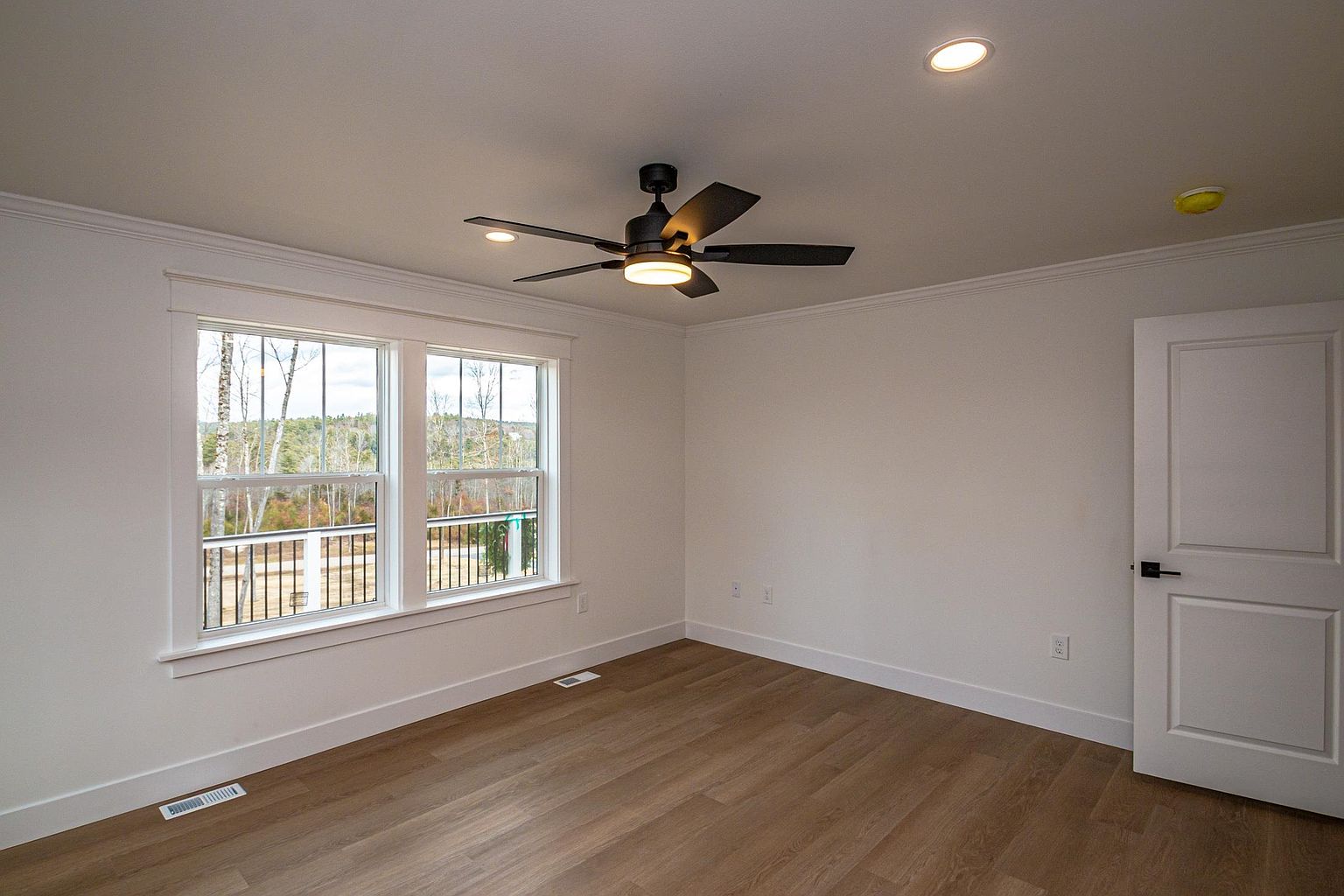 This bright and minimalist bedroom features light wood-look flooring, crisp white walls, and a modern black ceiling fan with integrated lighting. A large double window provides a view of the outdoor landscape, while a clean white door with black hardware adds a contemporary touch. The room is unfurnished, offering a blank canvas that emphasizes the home's neutral aesthetic and clean lines.