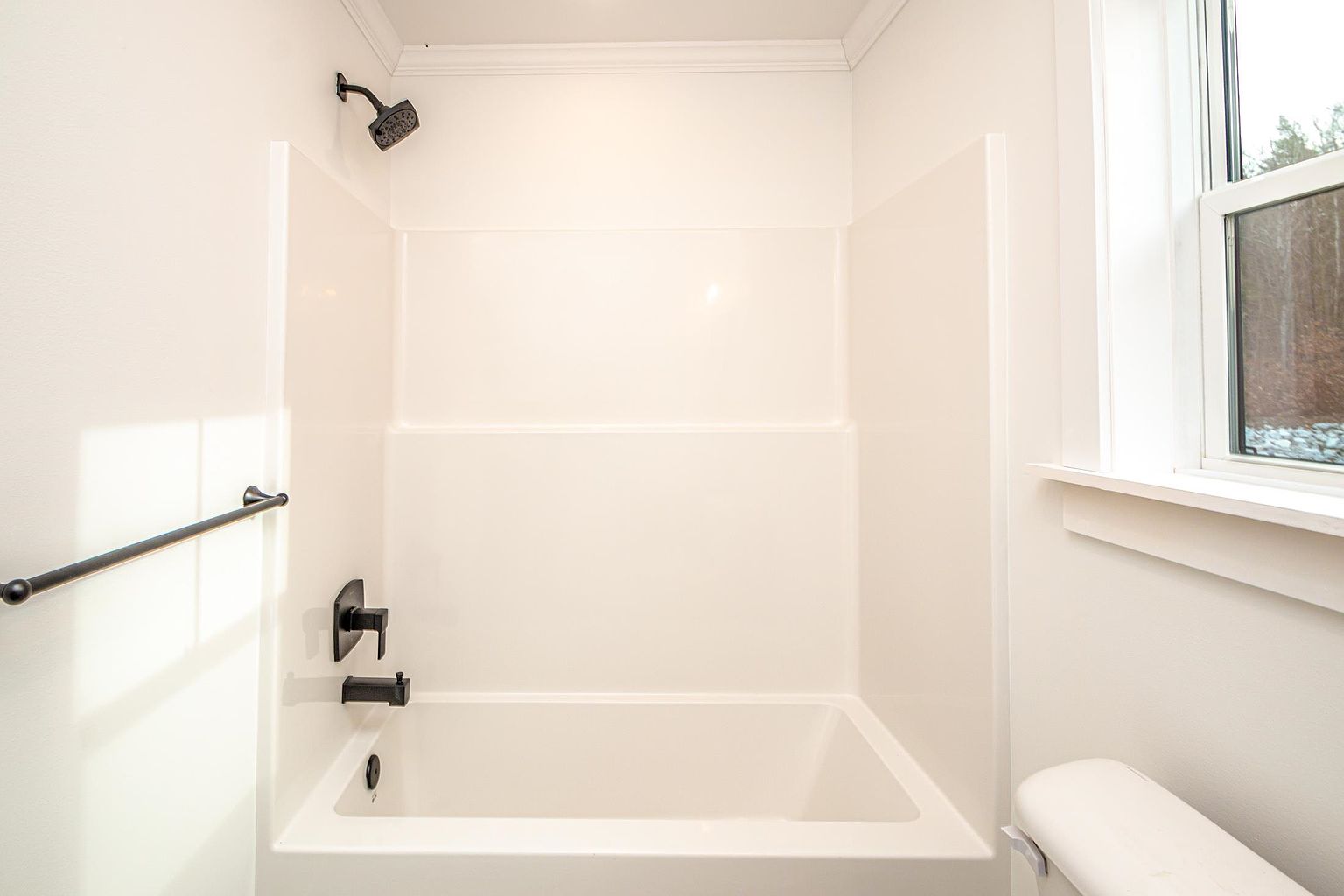This clean and minimalist bathroom features a white acrylic bathtub and shower surround, complemented by modern matte black fixtures. A simple grab bar is mounted on the side wall, and a window to the right allows for natural light to brighten the space. The neutral color palette and straightforward design create a functional and tidy appearance.