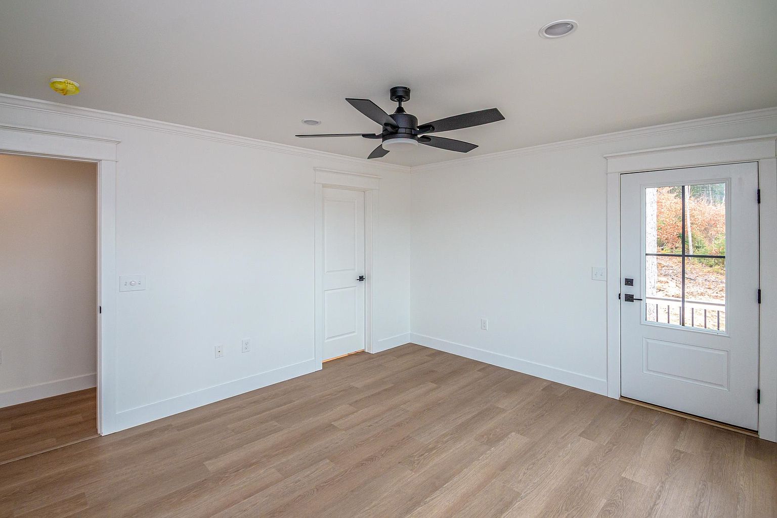 This bright and minimalist room features light wood-look flooring, crisp white walls, and a modern black ceiling fan. The space includes a white interior door, a glass-paned exterior door leading to the outdoors, and recessed lighting, creating a clean and versatile atmosphere. The perspective is a wide-angle shot from the corner of the room, emphasizing the open floor plan and neutral aesthetic.