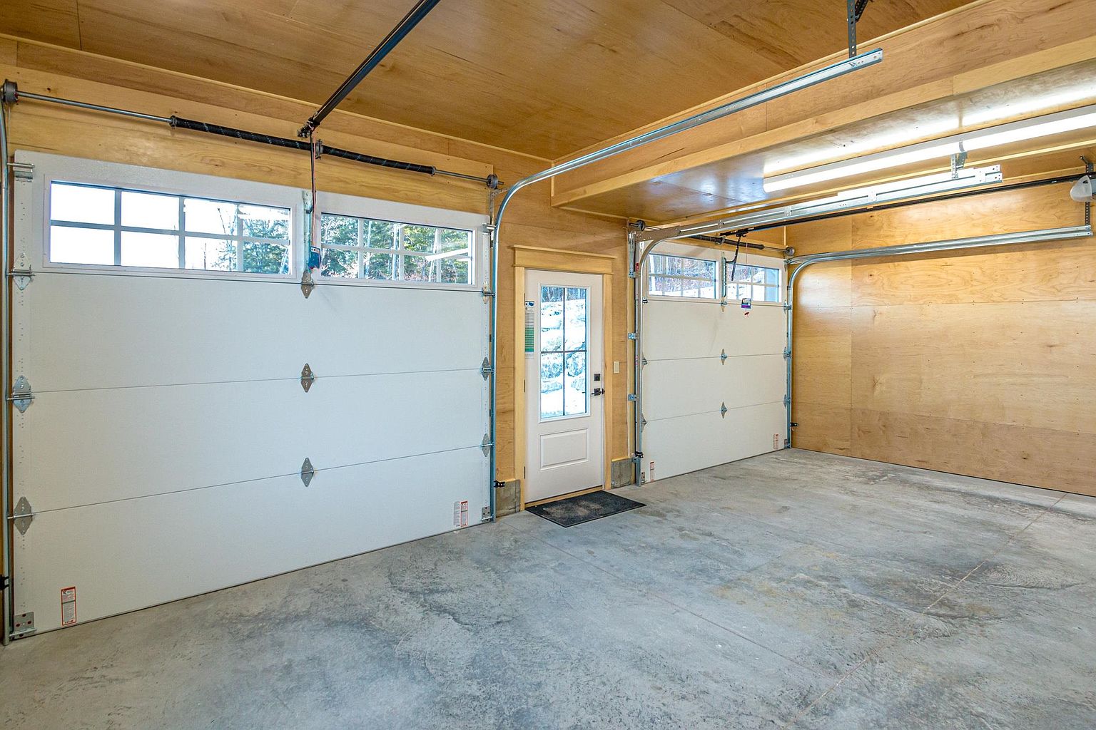 This image showcases a clean, well-maintained two-car garage interior featuring light-toned plywood walls and a smooth concrete floor. The space includes two white sectional garage doors with upper window panels and a central pedestrian door, all illuminated by bright overhead fluorescent lighting. The perspective is from the center of the garage, highlighting the organized, functional layout and the warm, wood-paneled aesthetic.