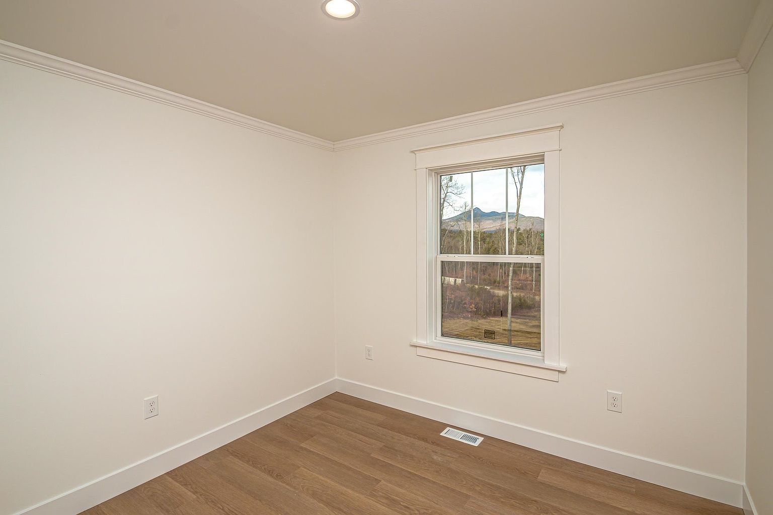 This bright, minimalist bedroom features neutral off-white walls, crown molding, and warm-toned wood-look flooring. A single window serves as the focal point, offering a scenic view of distant mountains and a wooded landscape. The room is clean and unfurnished, presenting a versatile blank canvas for potential buyers.