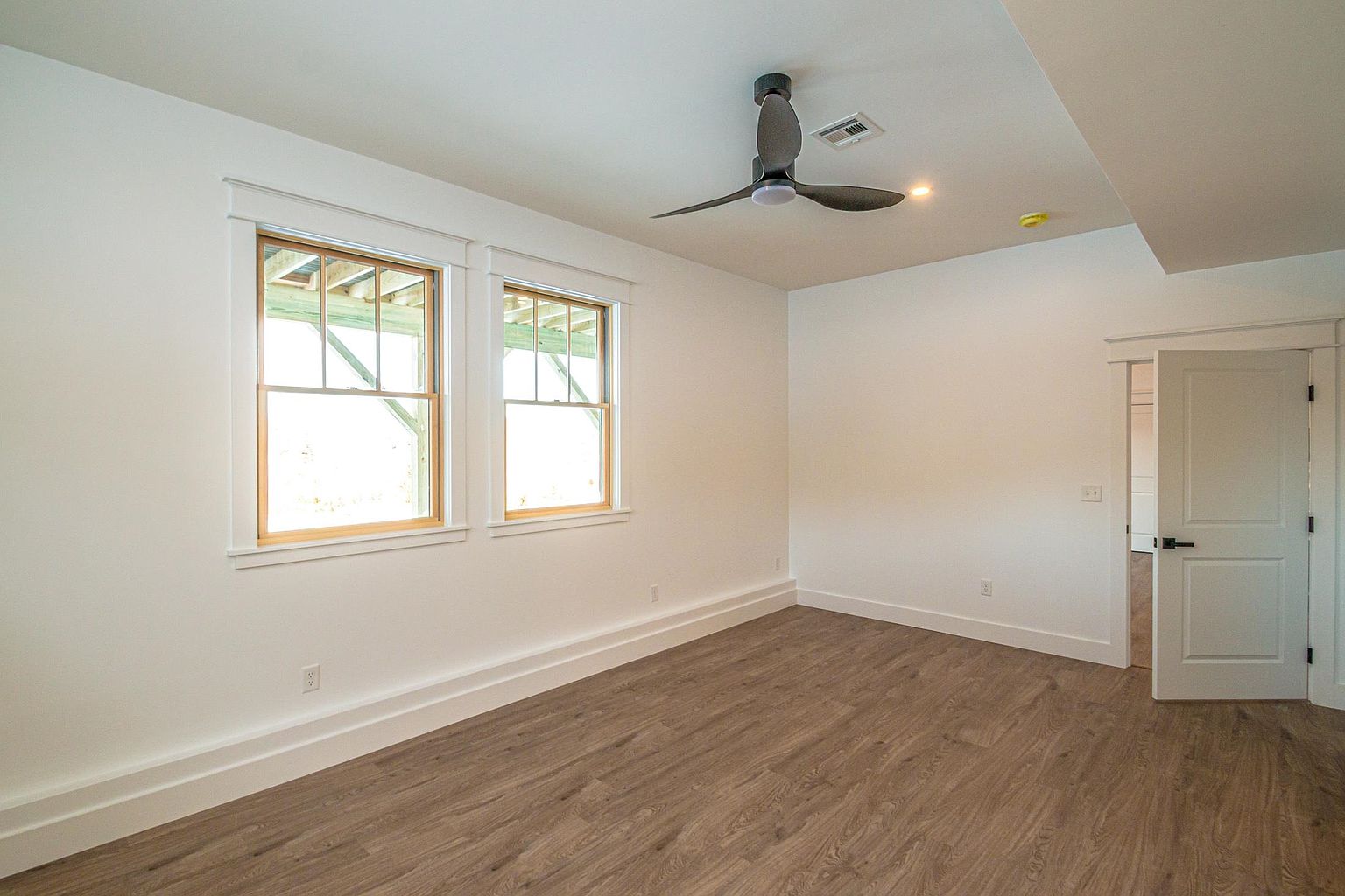 This bright and airy bedroom features neutral white walls, light wood-look flooring, and a modern ceiling fan. Two large windows with classic trim allow for ample natural light, while a doorway leads into an adjacent space. The room offers a clean, blank canvas aesthetic suitable for a guest bedroom or home office.