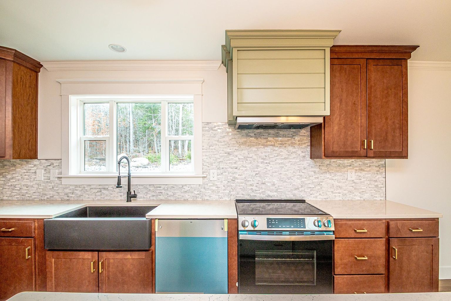 This modern kitchen features warm-toned wooden cabinetry paired with a light-colored quartz countertop and a textured mosaic tile backsplash. A prominent farmhouse-style sink sits beneath a large window, while a custom-built sage green range hood serves as a focal point above the electric stove. The space is well-lit and organized, offering a clean, functional, and inviting atmosphere.