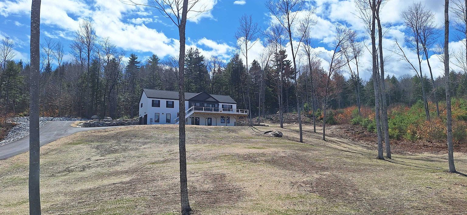 This wide-angle shot captures a modern, two-story home nestled in a serene, wooded landscape. The house features a prominent elevated deck, a paved driveway, and a clean, contemporary aesthetic that contrasts beautifully with the surrounding forest. The expansive, gently sloping lawn in the foreground provides a sense of privacy and space, making it an ideal setting for outdoor living.