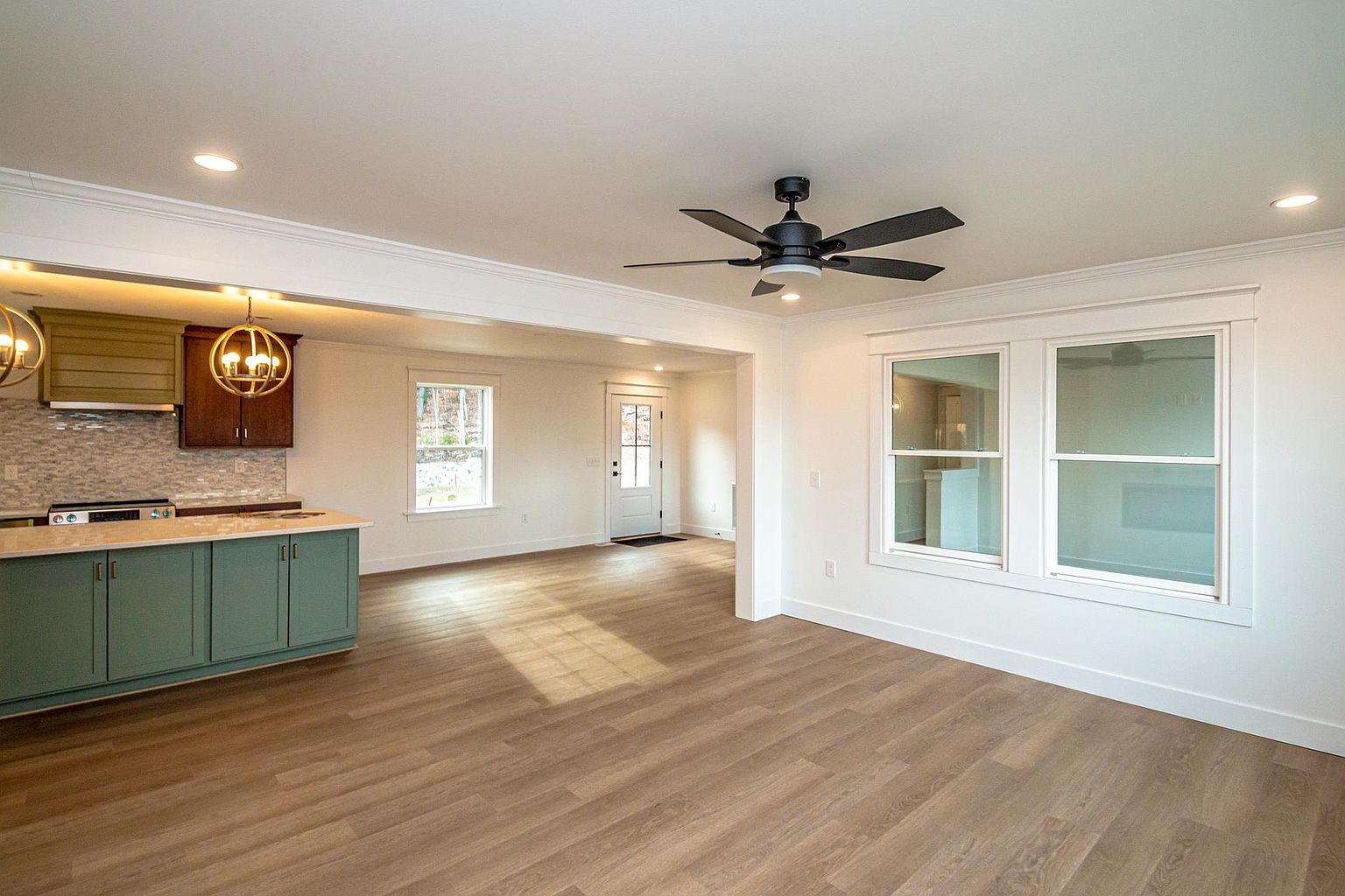 This open-concept space seamlessly blends a modern kitchen with a living area, featuring a sage green kitchen island and dark wood cabinetry. The room is brightened by recessed lighting, a stylish globe pendant light, and a ceiling fan, all set against neutral white walls and light wood-look flooring. The perspective captures the flow from the kitchen into the entryway, highlighting the home's airy and inviting layout.