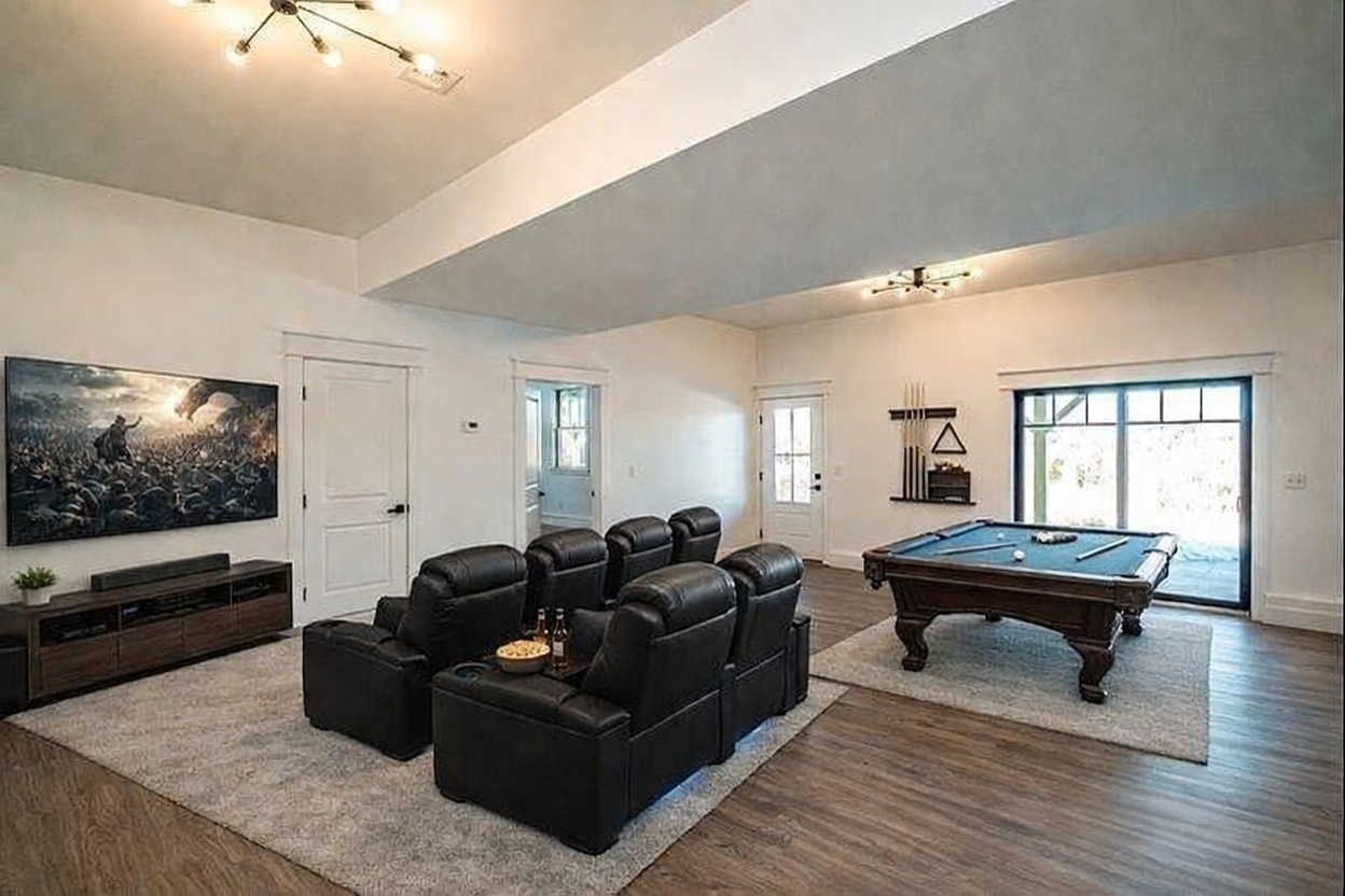 This spacious, open-concept basement features a dedicated home theater area with plush black leather recliners and a large wall-mounted television, complemented by a classic pool table in the adjacent space. The room is finished with modern wood-look flooring, neutral white walls, and contemporary lighting fixtures, creating a versatile entertainment hub. Large sliding glass doors provide natural light and access to the outdoors, enhancing the room's airy and inviting atmosphere.