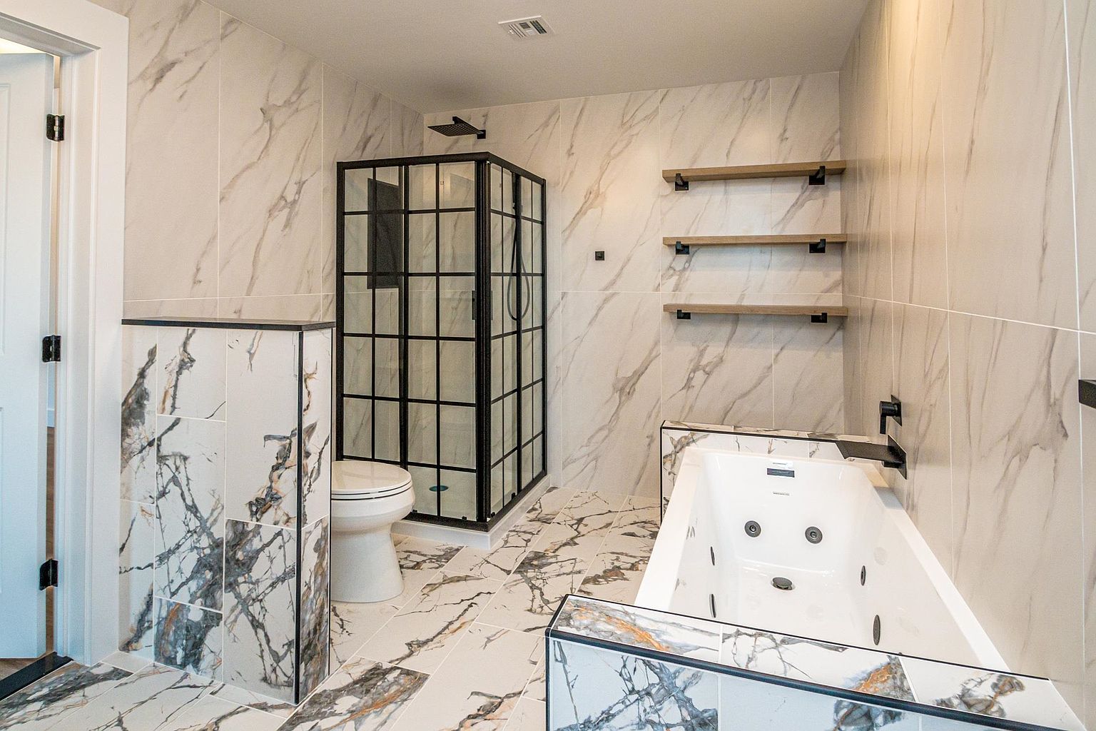 This luxurious primary bathroom features a striking modern design with bold, high-contrast marble tile flooring and wall accents. The space includes a sleek, black-framed glass shower enclosure, a deep jetted soaking tub, and minimalist floating wooden shelves, creating a sophisticated and spa-like atmosphere. The wide-angle perspective highlights the clean lines and premium finishes, emphasizing the contemporary aesthetic of the room.