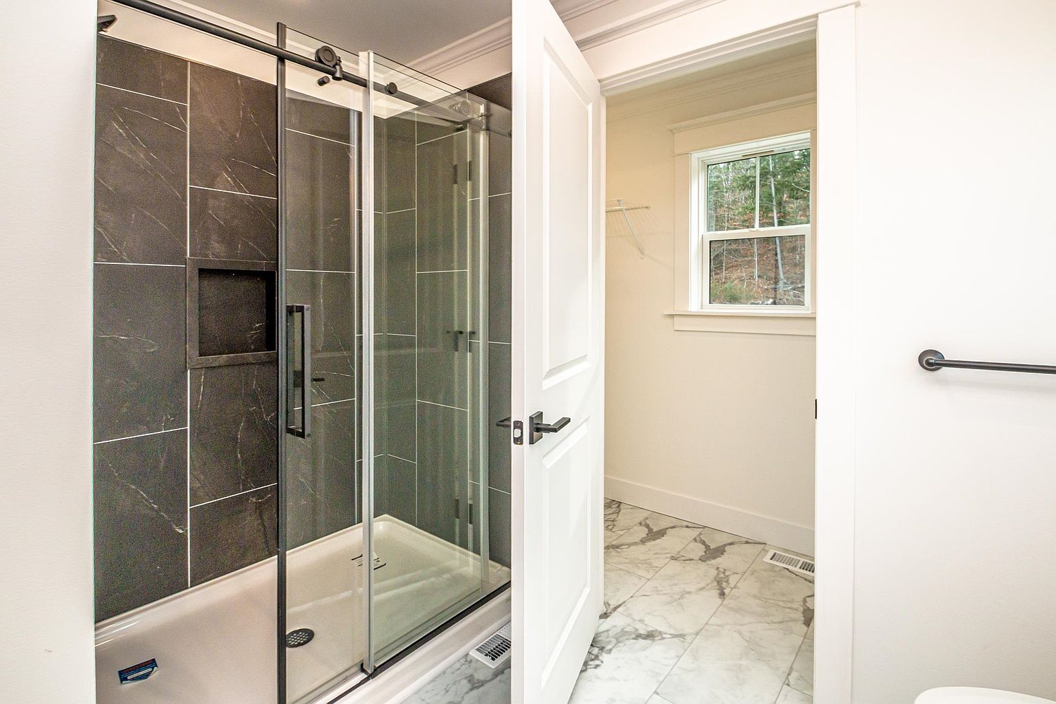This modern bathroom features a sleek glass-enclosed shower with dark, marble-patterned tile walls and a built-in niche. The space transitions into an adjacent walk-in closet through an open white door, showcasing a cohesive design with light marble-look floor tiles. The perspective captures the clean lines and contemporary finishes, highlighting the functional layout of the suite.