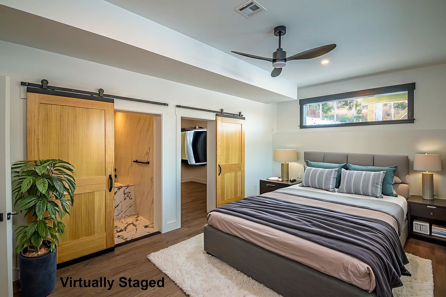 This modern primary bedroom features a neutral color palette with a plush gray upholstered bed and a soft white area rug. The room is accented by two natural wood sliding barn doors that lead to an en-suite bathroom and a walk-in closet, while a sleek ceiling fan and horizontal window add contemporary flair. The space feels warm and inviting, enhanced by the soft glow of bedside lamps and a potted plant in the corner.