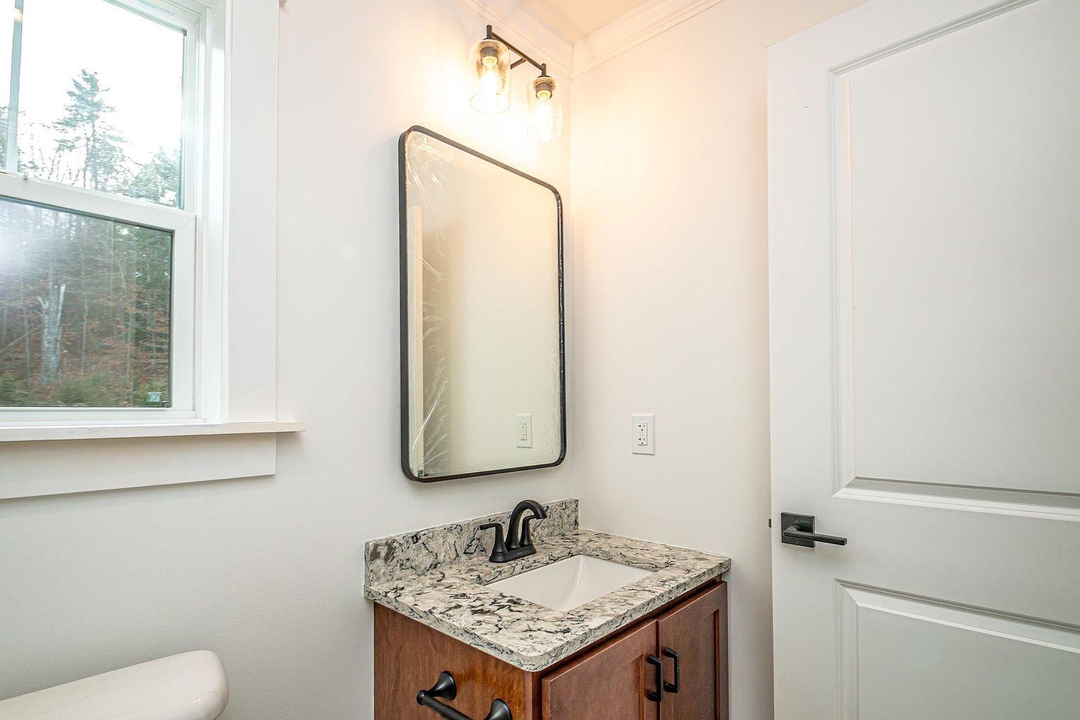 This compact, modern bathroom features a vanity with a granite countertop, a rectangular undermount sink, and a dark-finished faucet. A sleek, rectangular mirror with a dark frame hangs above the vanity, complemented by a dual-light fixture with Edison-style bulbs. The space is finished with neutral walls and a white door, creating a clean and functional aesthetic.
