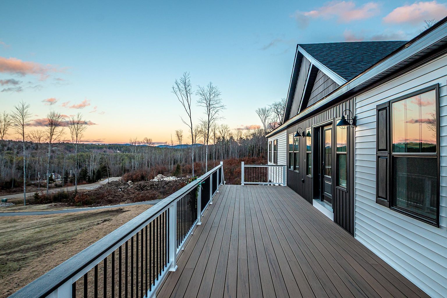 This image showcases a spacious, modern deck featuring composite wood-look decking and a sleek black metal railing that overlooks a scenic, rolling landscape at sunset. The home's exterior is finished with a combination of white horizontal siding and dark vertical board-and-batten accents, complemented by stylish wall-mounted light fixtures. The perspective captures the expansive outdoor living space, emphasizing the peaceful, elevated view of the surrounding forest and hills.