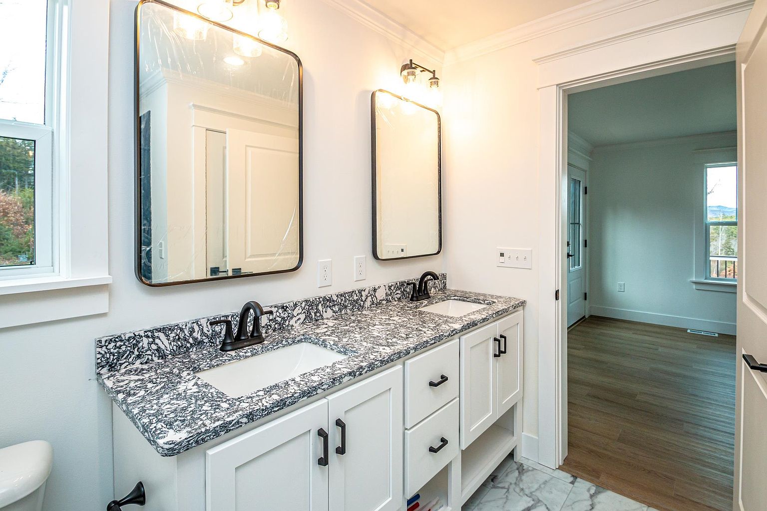 This bright and modern bathroom features a double vanity with a striking black-and-white speckled granite countertop and white shaker-style cabinetry. Two rectangular mirrors with dark frames are mounted above the sinks, complemented by sleek black hardware and fixtures. The space is well-lit and leads into an adjacent room with light wood flooring, creating a clean and cohesive aesthetic.