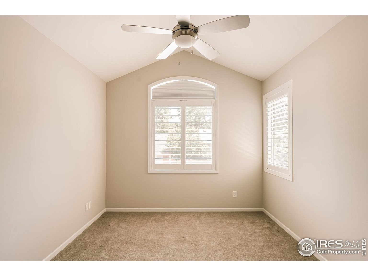 The image showcases a well-lit bedroom with neutral-toned walls and carpet. A window with white shutters provides natural light, and a ceiling fan adds to the room's comfort. The room appears clean and ready for occupancy, suitable for a guest or child's bedroom.