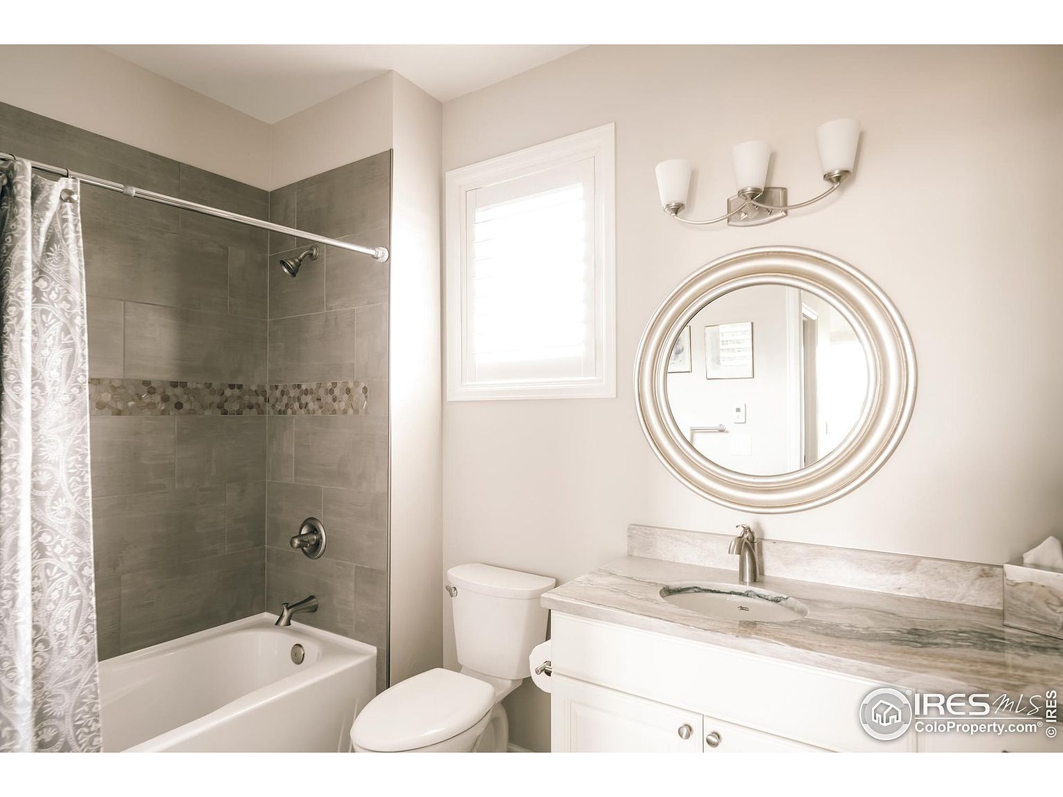 This is a well-lit bathroom featuring a bathtub with gray tiled walls and a decorative mosaic strip, complemented by a shower curtain. A white toilet and vanity with a marble countertop and round mirror are also visible. The overall impression is clean and modern.