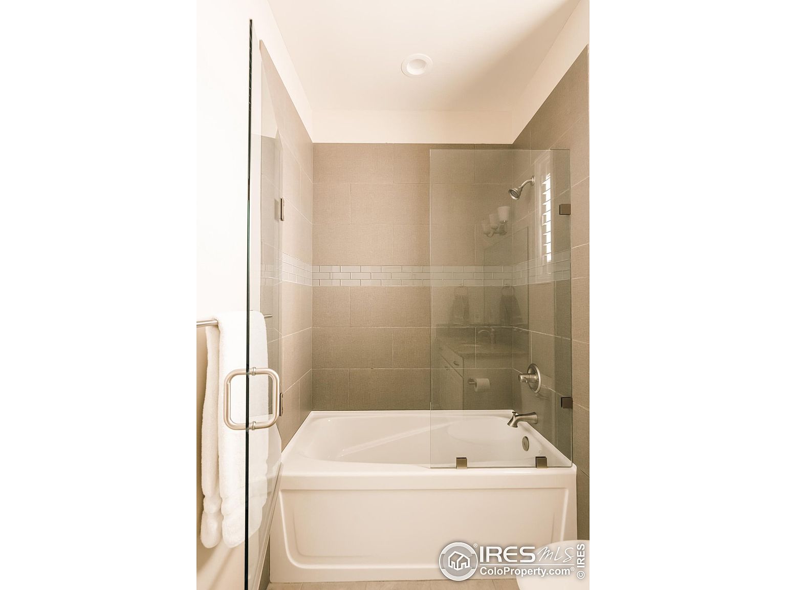 This is a well-lit bathroom featuring a bathtub with a partial glass enclosure. The walls are tiled in a neutral tone, complemented by a horizontal accent strip. A towel hangs on the glass door, and the overall impression is clean and modern.