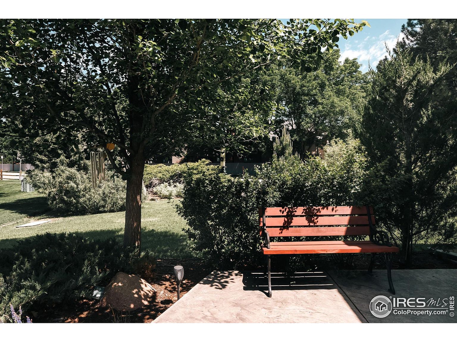 This image showcases a serene yard or garden setting, featuring a wooden bench nestled among lush greenery. The scene is shaded by mature trees, creating a tranquil and inviting space. A stone pathway and well-maintained lawn add to the property's appeal, suggesting a peaceful outdoor retreat.