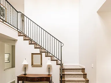 This interior shot highlights a staircase with carpeted steps and a wrought iron railing, leading to the upper level of the house. Below the stairs, a wooden console table decorated with lamps adds a touch of elegance. The wall's neutral color palette allows natural light to brighten the space.