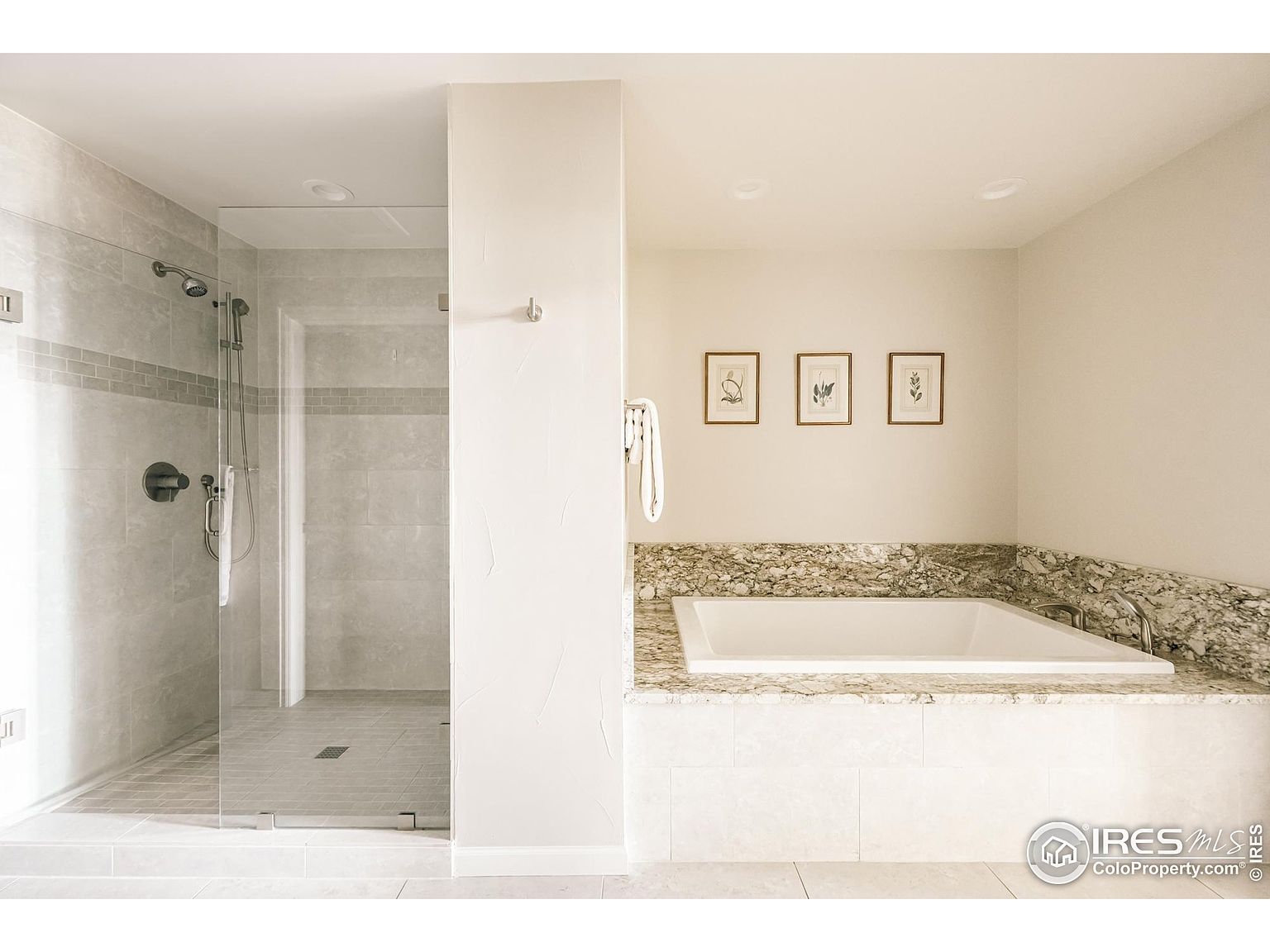 This is a well-lit primary bathroom featuring a glass-enclosed shower with gray tiling and a separate soaking tub with a granite surround. Three framed botanical prints adorn the wall above the tub, adding a touch of elegance. The overall impression is clean, modern, and luxurious, highlighting the spa-like atmosphere.