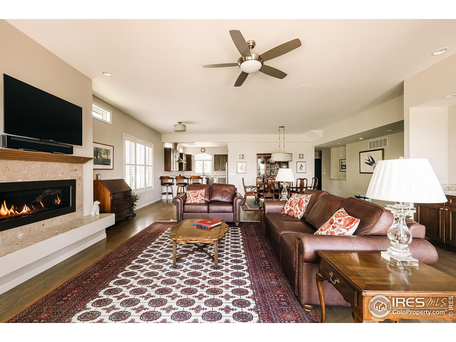This is an interior shot of a living room featuring a fireplace with a mounted television above it. The room is furnished with leather sofas, a patterned rug, and wooden side tables. The space is open and flows into the dining area, creating a welcoming and comfortable atmosphere.