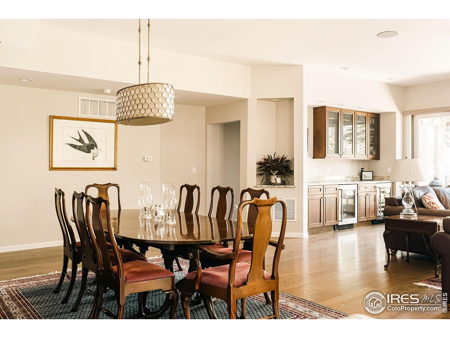 This is an interior shot of a dining room featuring a large, round wooden table surrounded by eight chairs with red cushions. A decorative rug lies beneath the table, and a modern chandelier hangs above. The room transitions into a bar area with wooden cabinets and a glimpse of a living room, creating an open and inviting space.