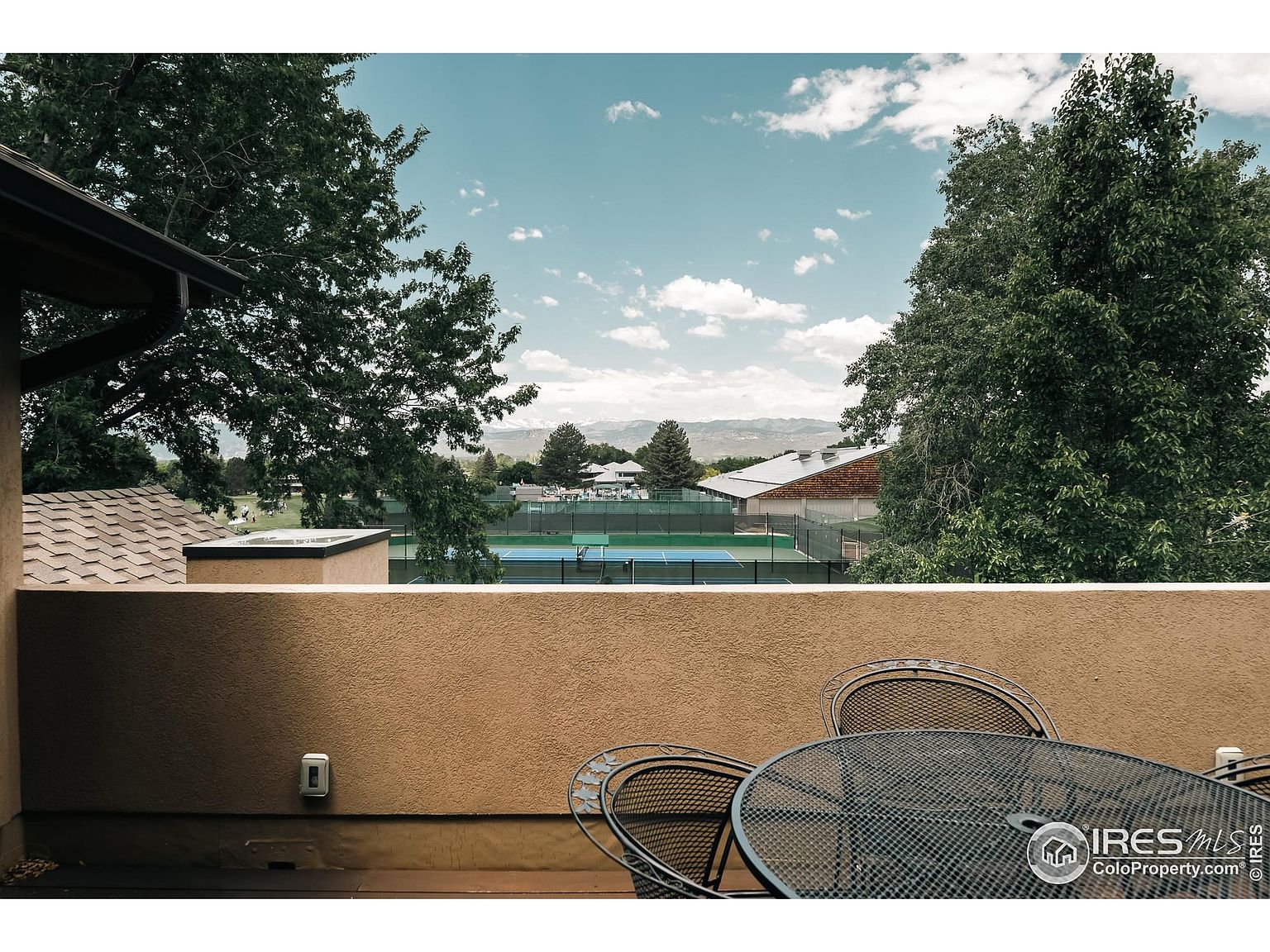 This image showcases a balcony or patio area with a view of tennis courts and distant mountains. The foreground features a metal patio table and chairs, suggesting a relaxing outdoor space. The overall impression is one of leisure and access to recreational amenities.