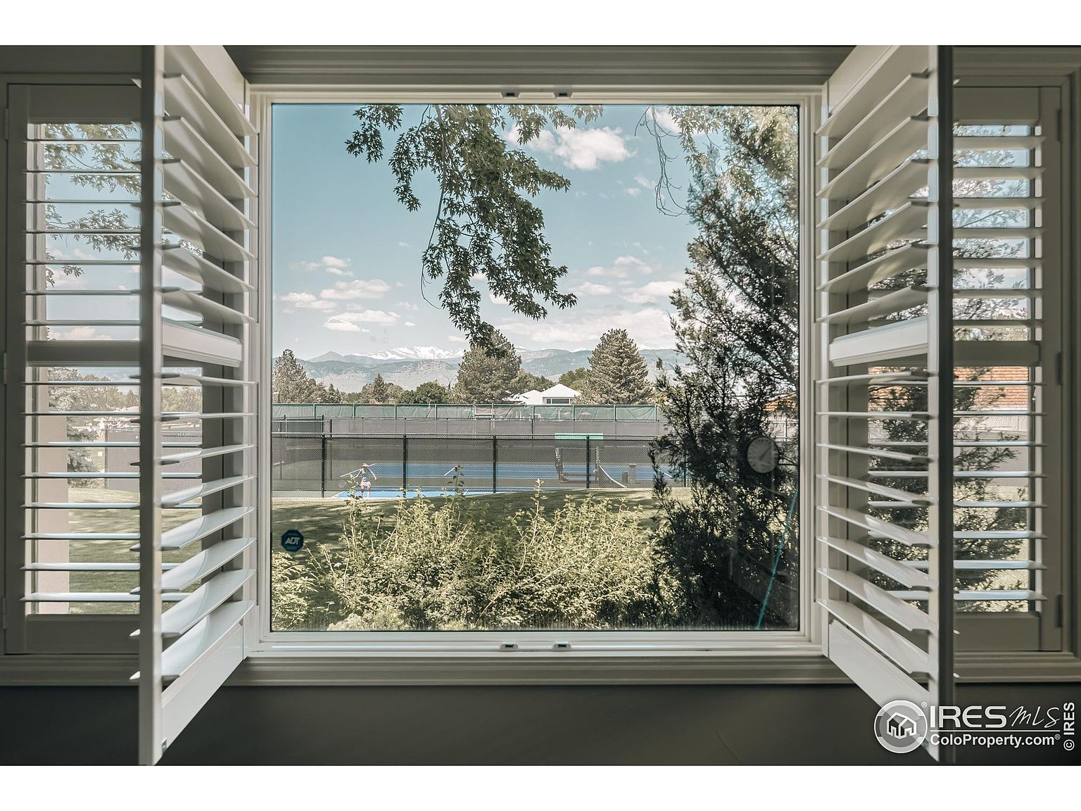 The image showcases a view from inside a room, looking out through an open window with white shutters. The window frames a scene of a tennis court, lush greenery, and distant mountains under a partly cloudy sky. The perspective is from inside the house, emphasizing the connection between the interior space and the outdoor recreational area.