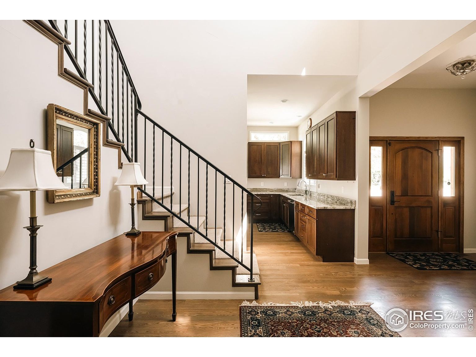 This interior shot showcases a welcoming entryway and staircase. A wooden console table with lamps sits against the wall, complemented by a framed mirror. The staircase features black iron railings and leads to the upper level, while a glimpse of the kitchen and a wooden front door with sidelights add depth to the scene. The hardwood flooring and area rug enhance the home's warmth and character.