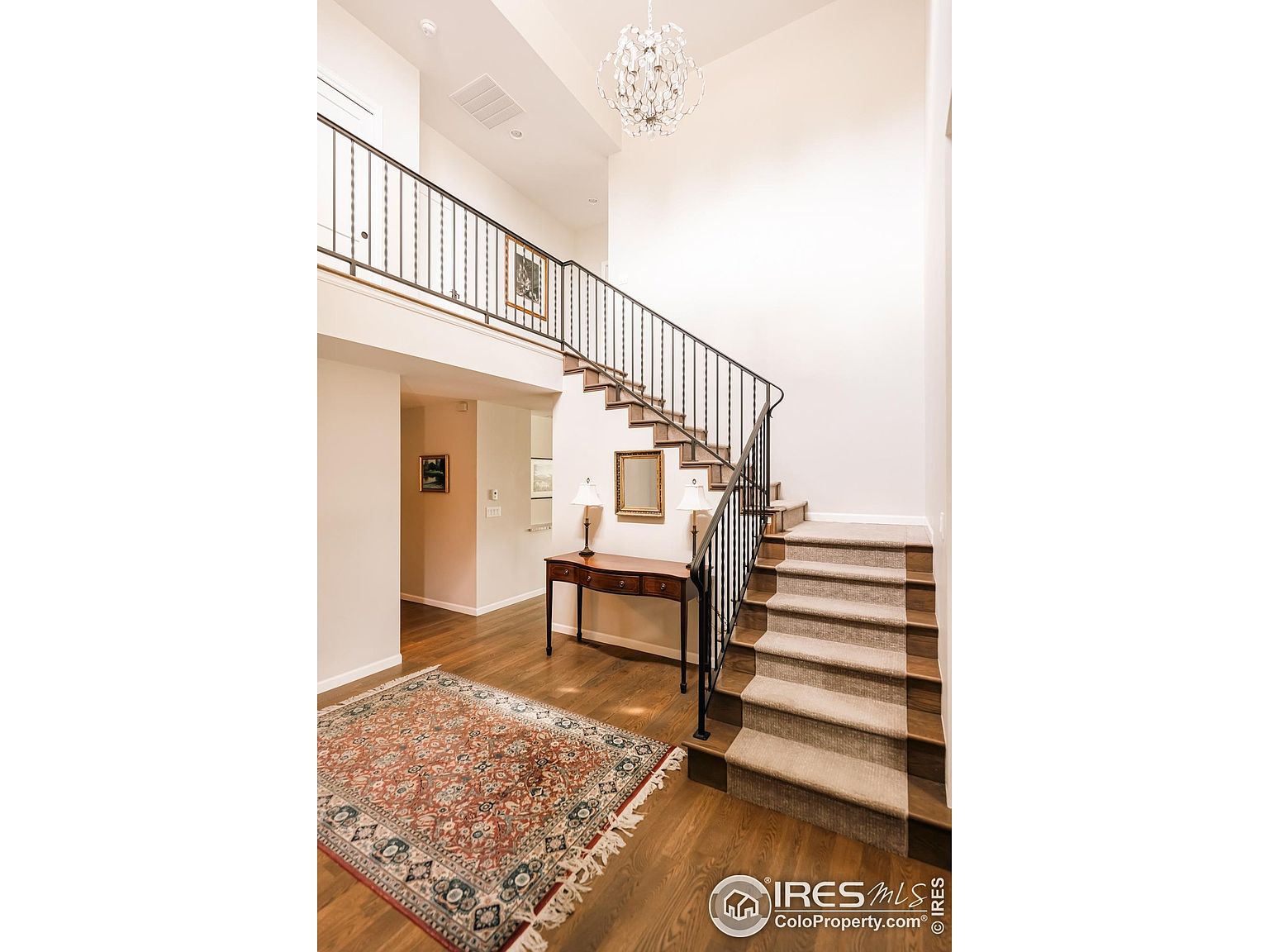 This is an interior shot of a hallway and staircase. The staircase is carpeted and has a dark metal railing. There is a table with lamps and a mirror in the hallway, and a decorative rug on the hardwood floor. The walls are painted in a neutral color, and a chandelier hangs from the ceiling.