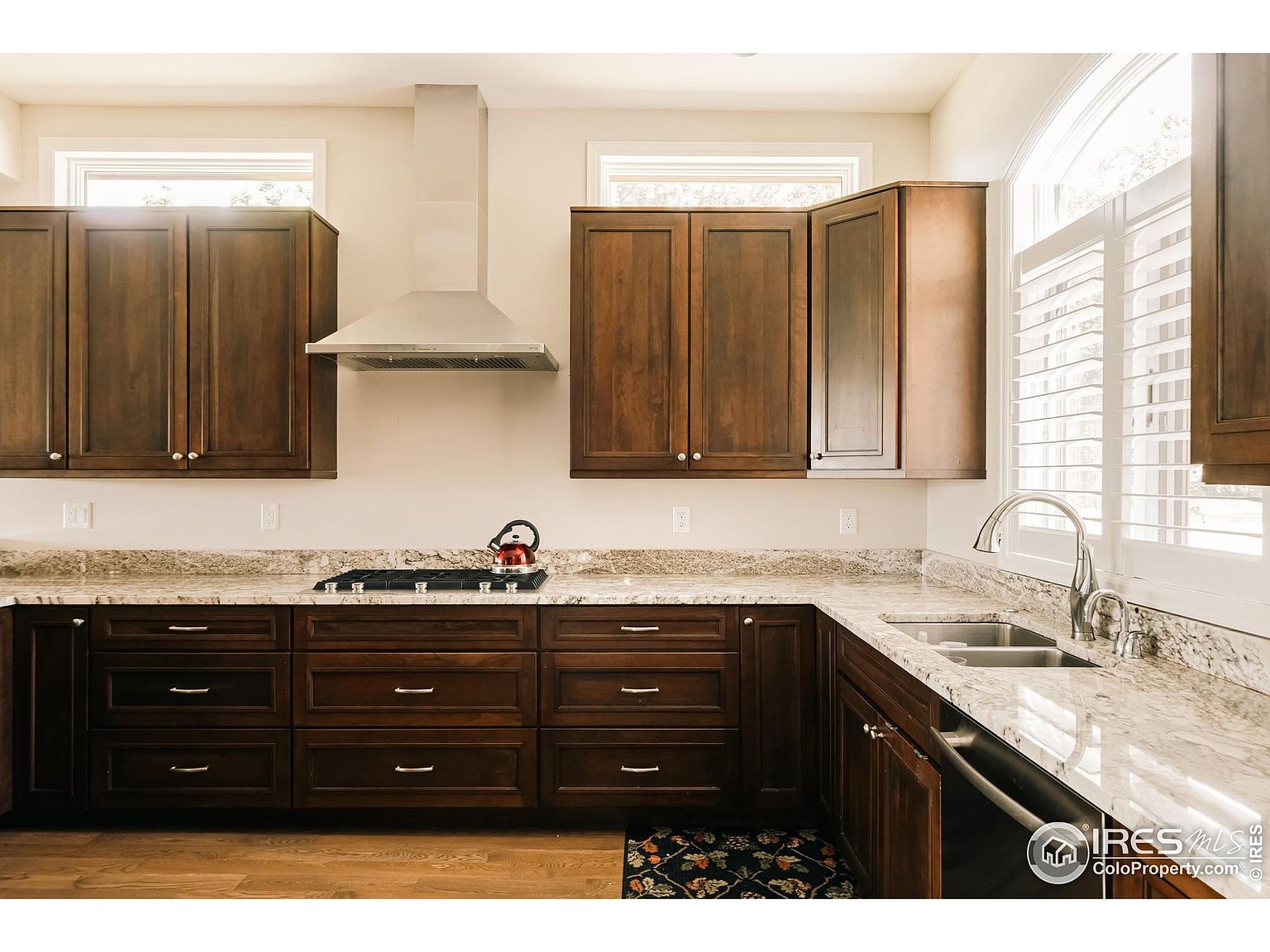This is a well-lit kitchen featuring dark wood cabinetry, granite countertops, and stainless steel appliances. A gas cooktop is visible on the countertop, and a stainless steel range hood is mounted above it. The kitchen also has a double sink and a window with white shutters, creating a warm and inviting atmosphere.