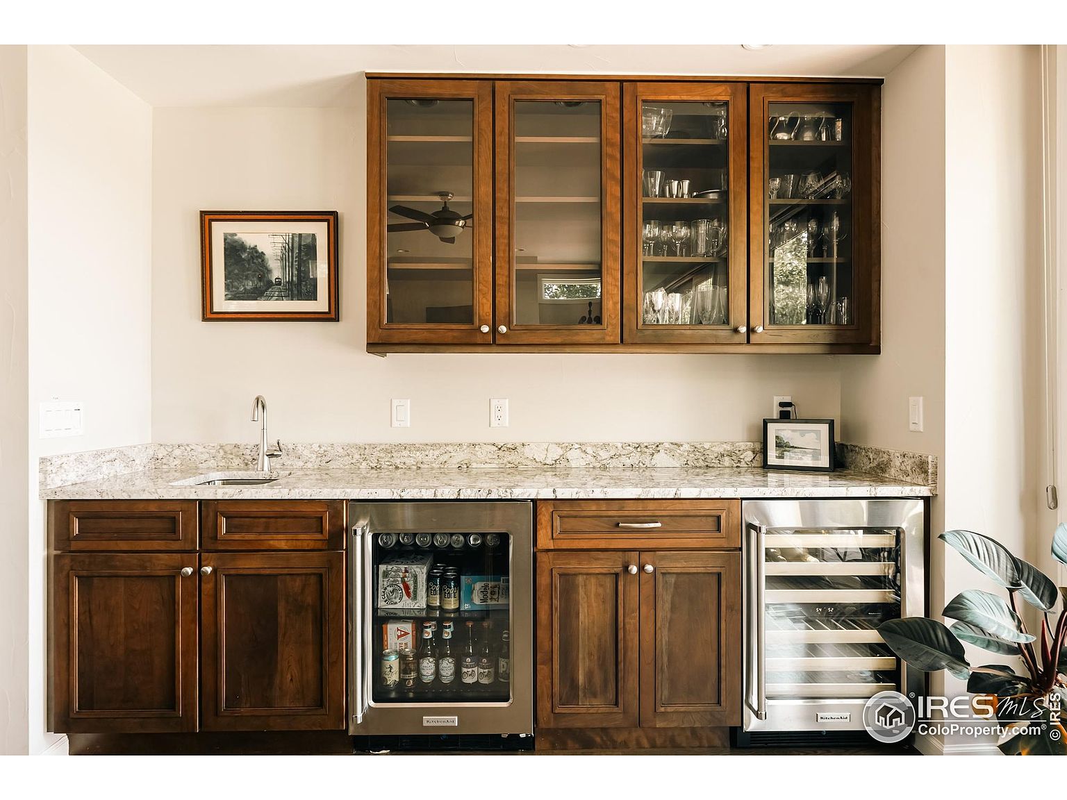 This interior shot showcases a well-appointed kitchen bar area with rich wood cabinetry, glass-fronted upper cabinets displaying glassware, and a light-colored granite countertop. The space includes a beverage refrigerator and a wine cooler, suggesting a focus on entertaining. A framed picture adds a touch of personality to the space.