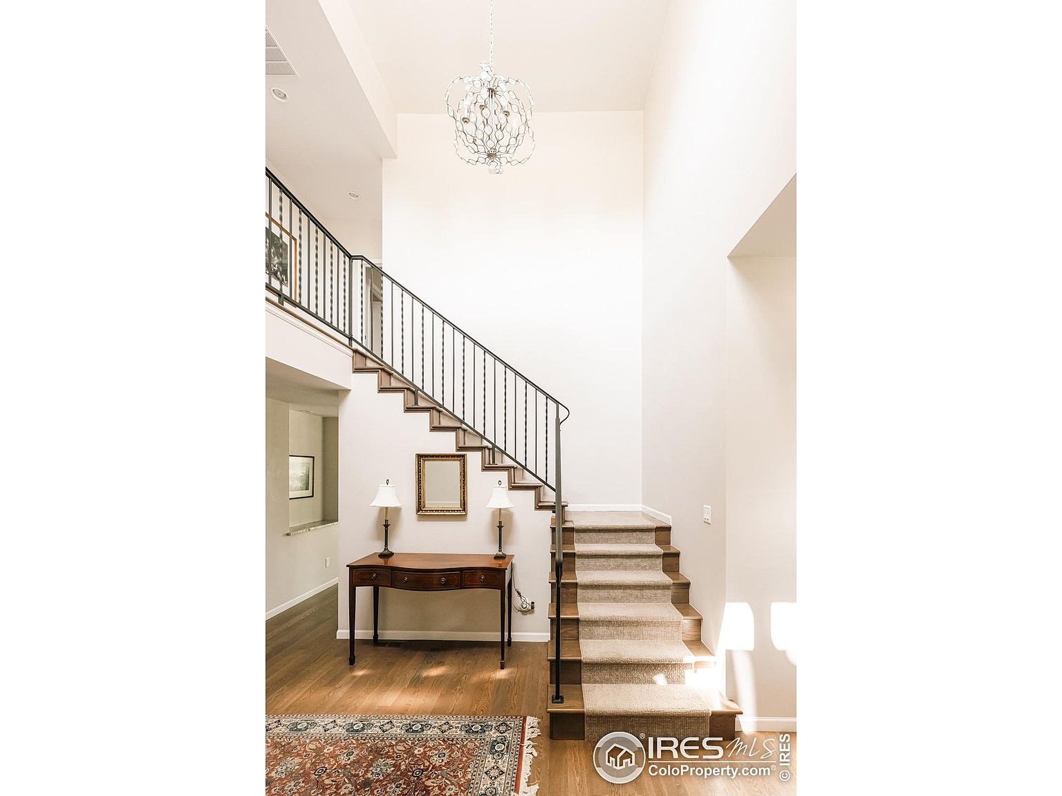 This interior shot showcases a grand hallway and staircase. The staircase features carpeted steps and a dark metal railing, leading to an upper level. A console table with lamps sits adjacent to the stairs, adding a touch of elegance to the space, while a chandelier hangs from the high ceiling, illuminating the area.